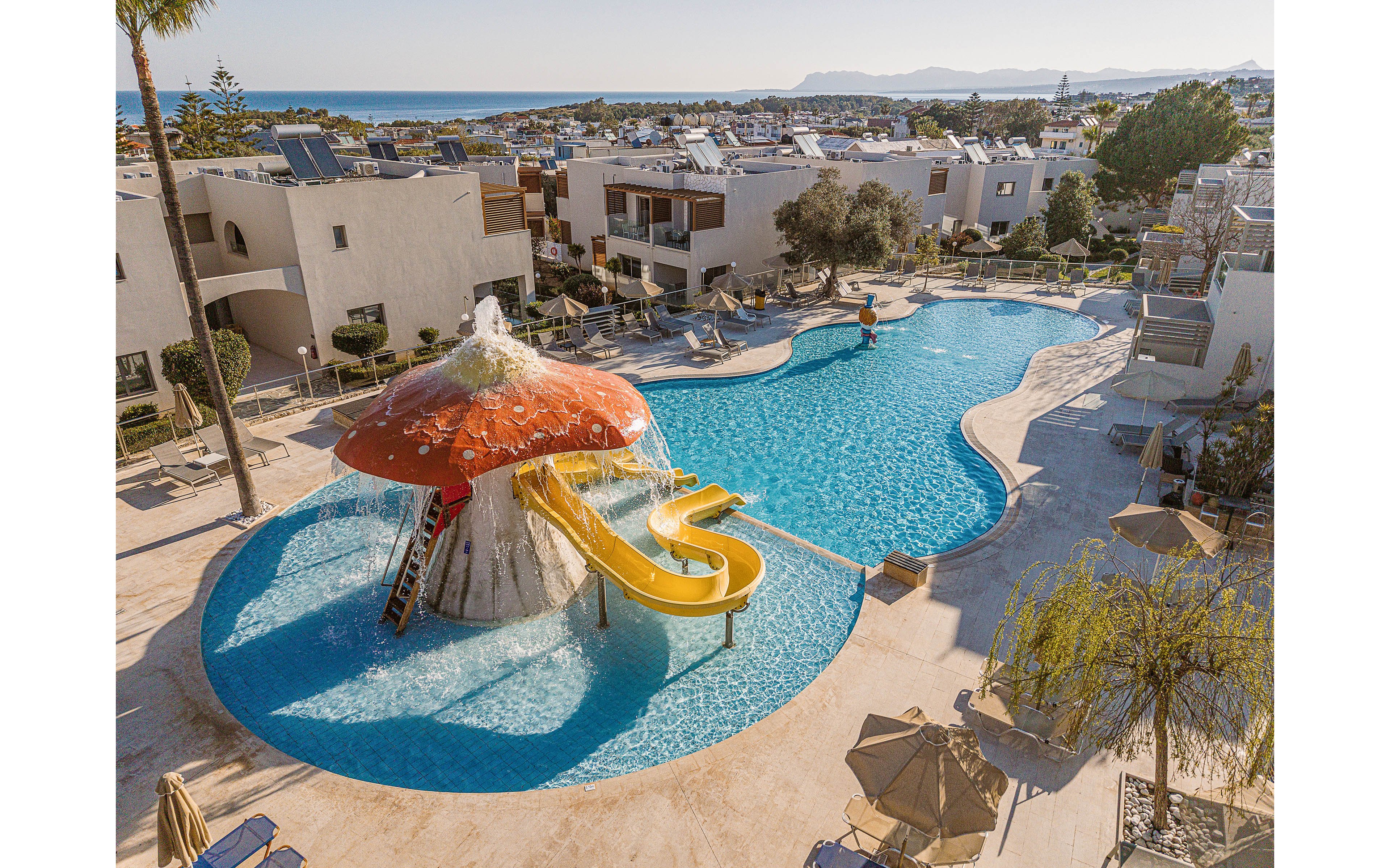 A large resort swimming pool features a playful mushroom-shaped water structure with a yellow slide. The pool is surrounded by sun loungers, umbrellas, and modern low-rise buildings. In the background, the coastline and blue sea are visible under a clear sky. The scene suggests a sunny vacation destination designed for family leisure and relaxation.