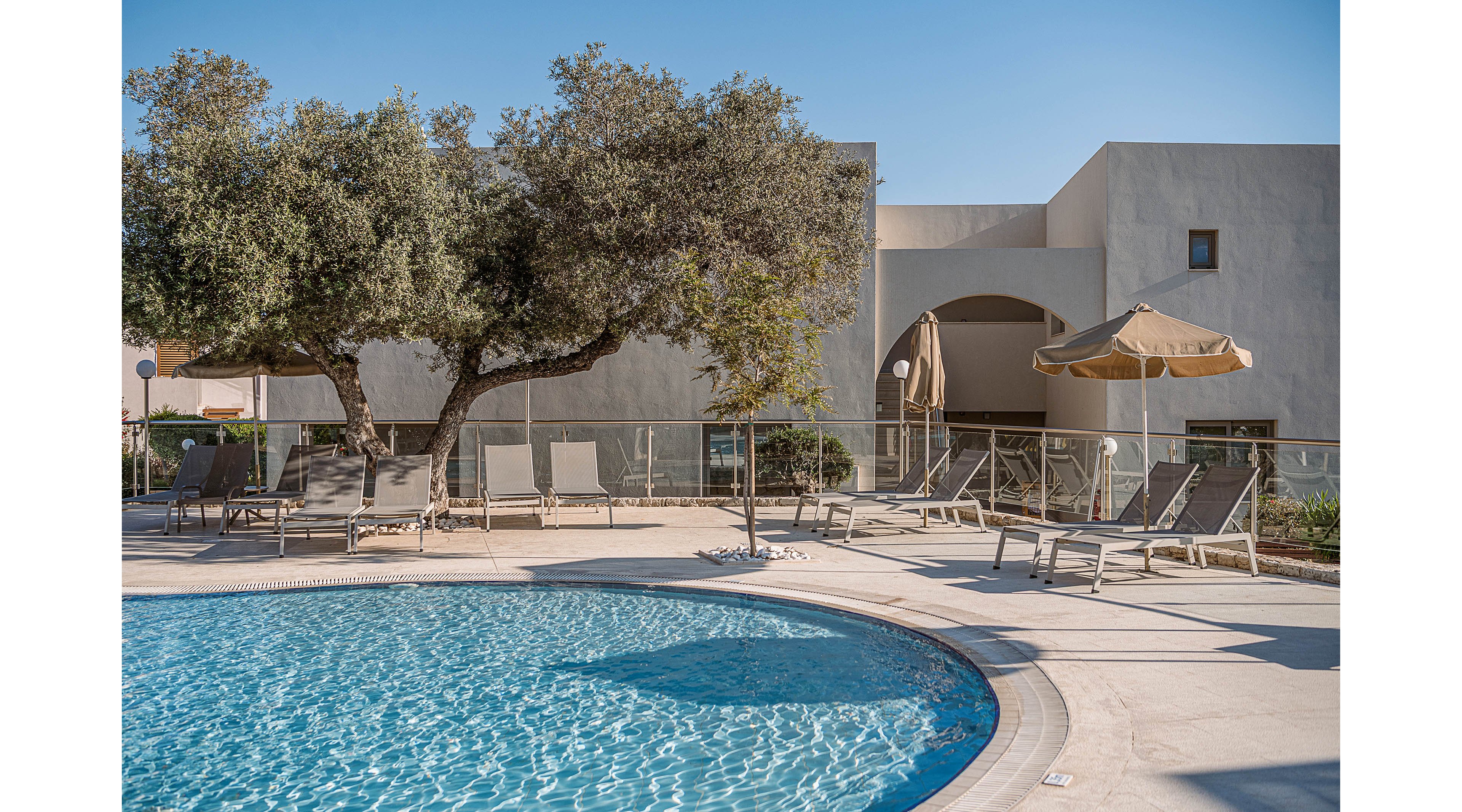 An outdoor swimming pool glistens under clear blue skies at a modern resort. Several lounge chairs and umbrellas are arranged around the poolside deck, inviting relaxation. A large tree provides natural shade near the seating area, contrasting with the clean lines of the light-colored building in the background. The scene feels calm and quiet, suggesting a peaceful vacation setting.