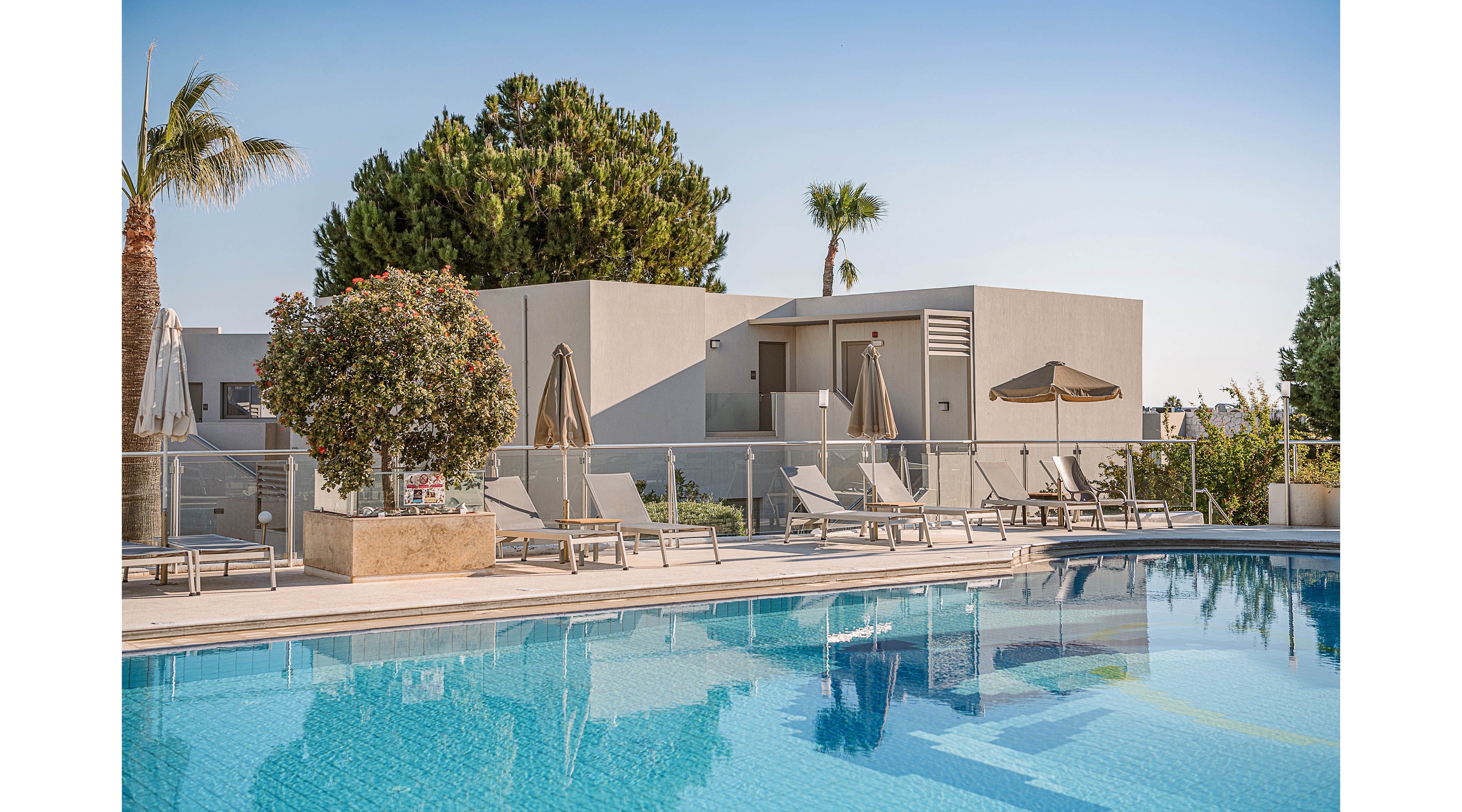 A clear blue swimming pool reflects a row of modern, minimalist resort buildings. Several sun loungers and closed umbrellas line the poolside terrace, suggesting a quiet and relaxing atmosphere. Palm trees and lush greenery add a touch of tropical nature to the contemporary architecture. The bright daylight and cloudless sky create a warm, inviting vacation setting.