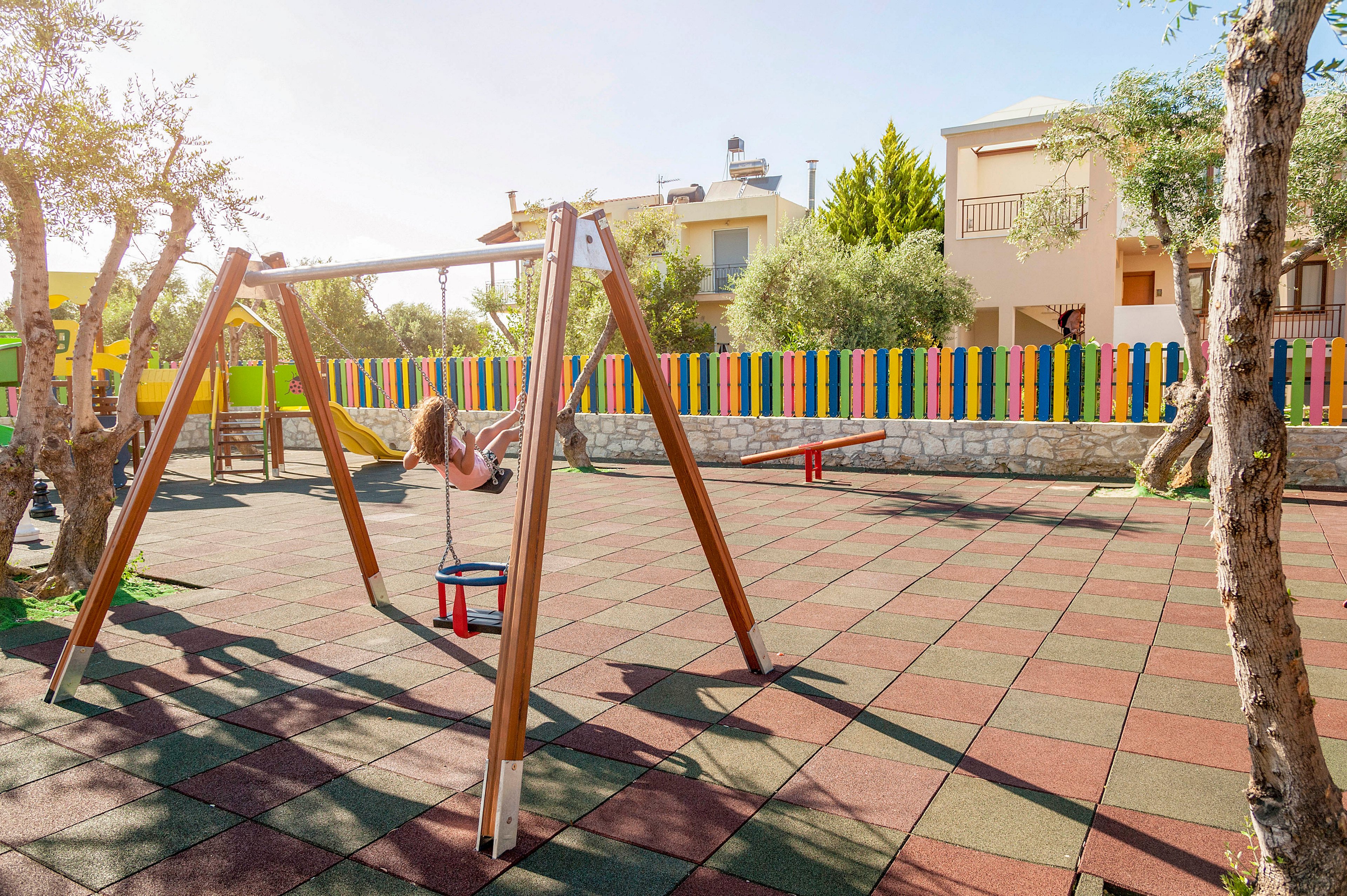 A child swings on a wooden swing set in an outdoor playground. The play area features colorful rubber tiles and several pieces of playground equipment, including a seesaw in the background. A bright, multicolored fence surrounds the space, with residential buildings and trees beyond it. The scene is lit by warm sunlight, creating a cheerful and inviting atmosphere.