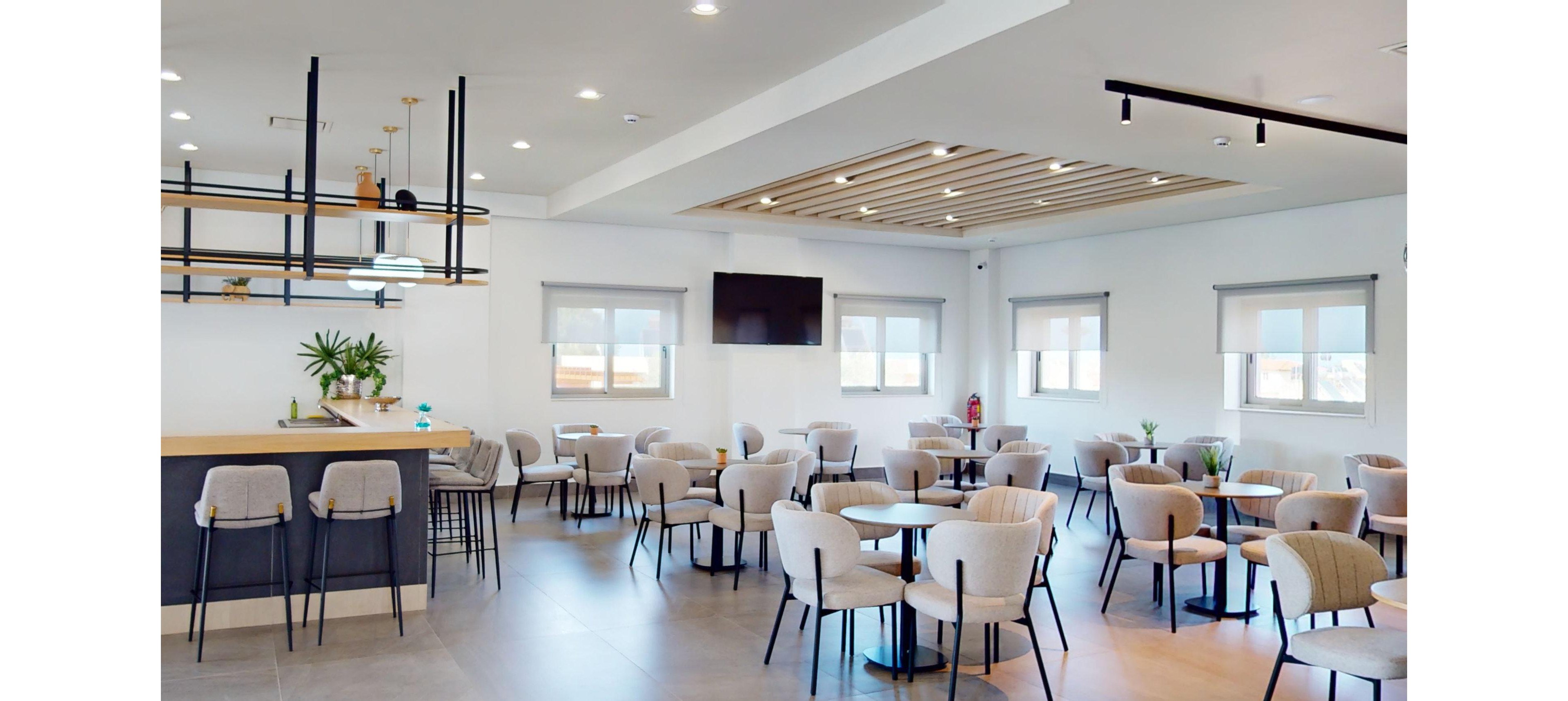 A modern cafeteria interior features multiple small round tables with upholstered chairs neatly arranged in an open space. A long counter with high stools and minimalist shelving stands on the left side of the room. Large windows with roller shades allow natural light to brighten the neutral-toned decor. A flat-screen television is mounted on the wall, and recessed ceiling lights add to the clean, contemporary atmosphere.