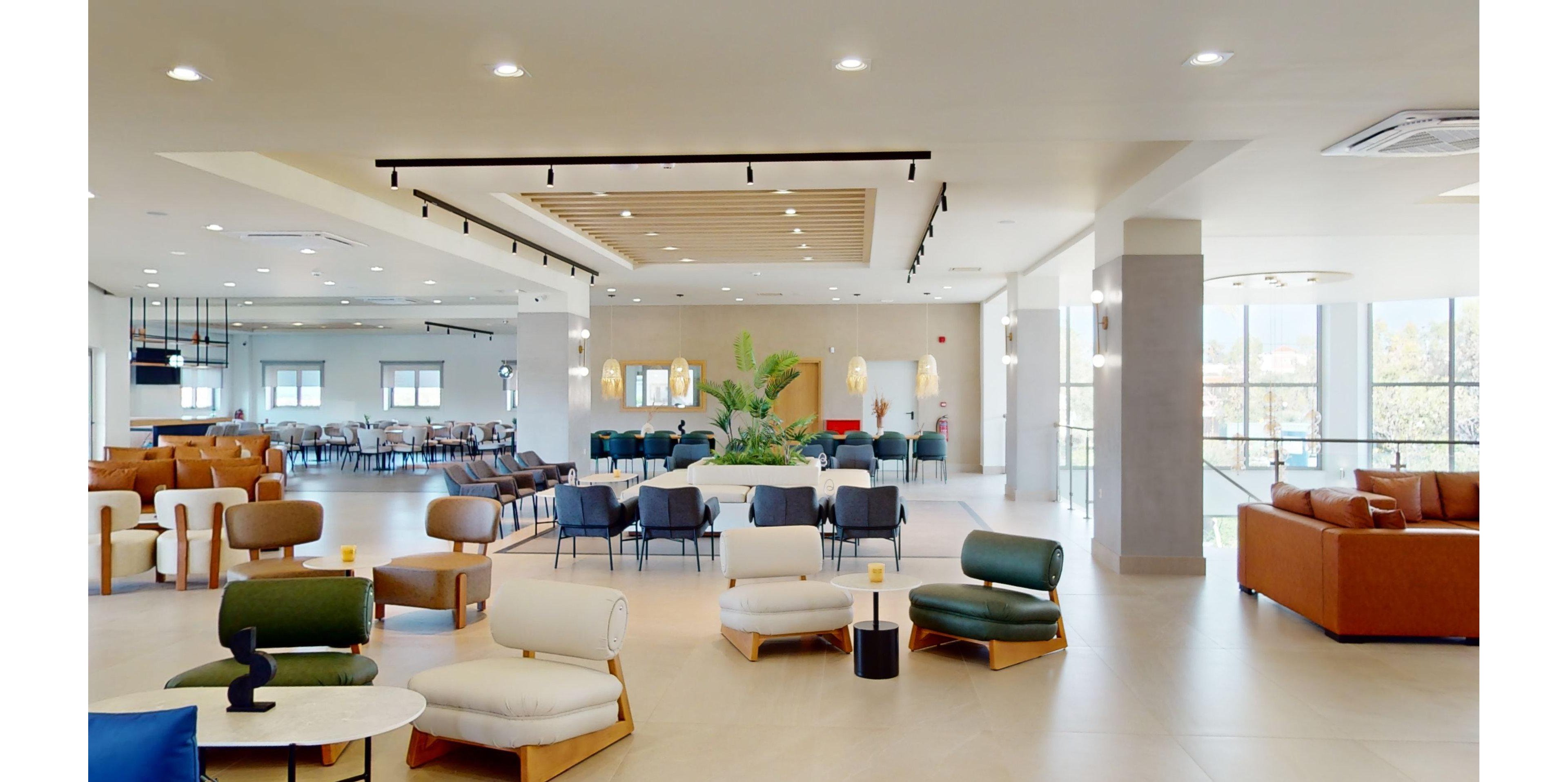 Spacious modern lobby features colorful contemporary lounge chairs and sofas arranged in conversational groupings. The setting appears to be a bright communal area with large windows and abundant natural light. A central planter with tall green plants adds a biophilic touch to the minimalist interior. Neutral floors and walls contrast with warm leather and fabric upholstery, creating a welcoming, flexible workspace or hospitality environment.