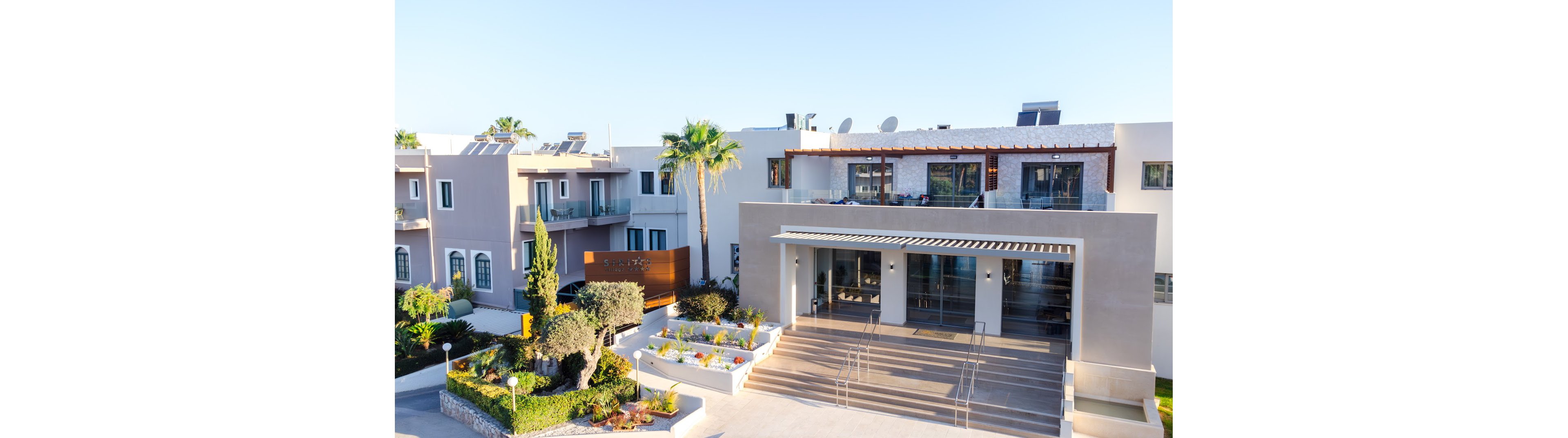 A modern hotel complex is shown from an elevated viewpoint in bright daylight. The building features clean lines, large glass entrances, and multiple balconies overlooking landscaped gardens. Palm trees, shrubs, and flower beds decorate the front courtyard and stepped walkway. The clear blue sky and strong sunlight create a warm, inviting atmosphere suitable for vacation or resort imagery.