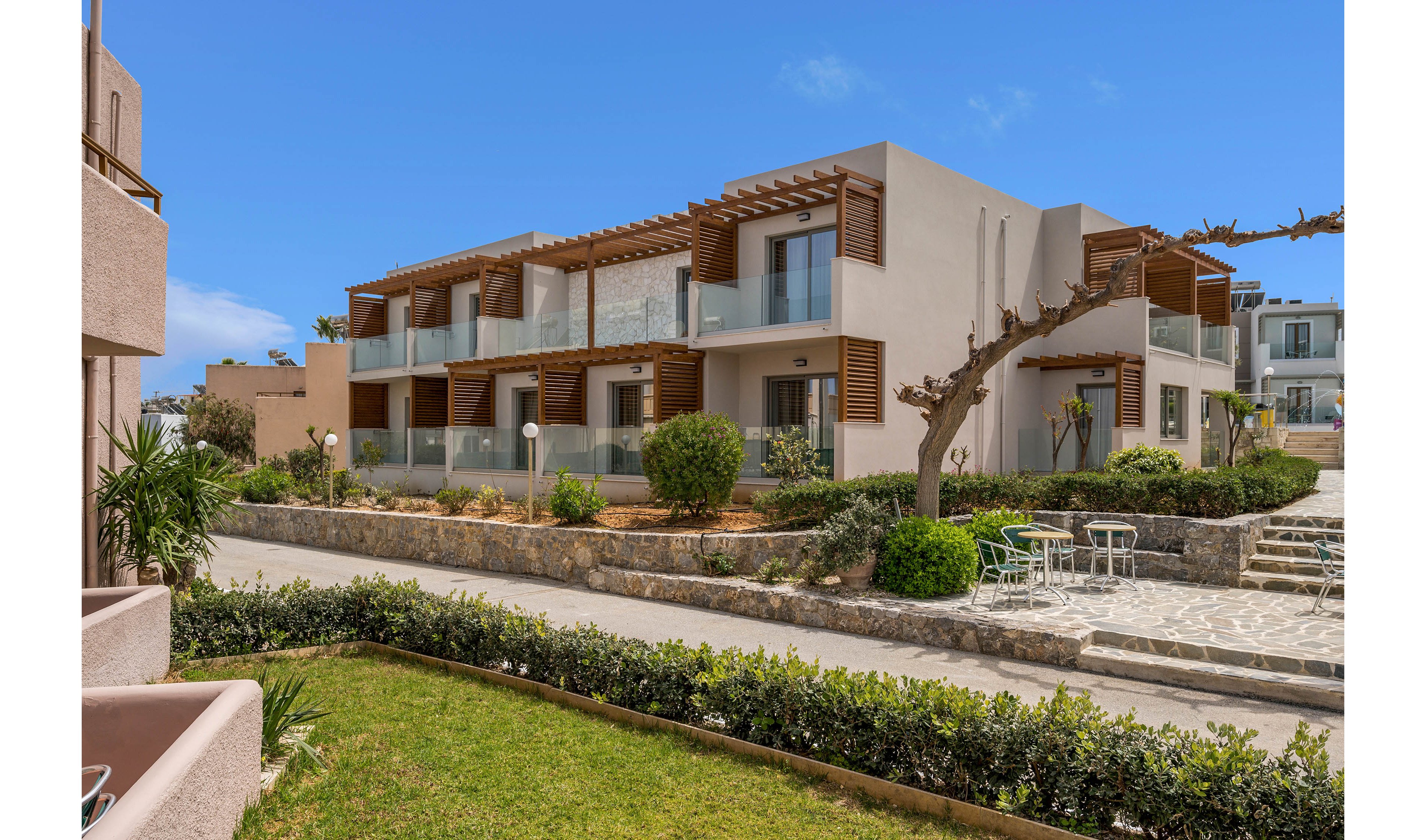 A modern two-story apartment complex features clean lines, glass balconies, and wooden pergolas. The buildings are surrounded by neatly landscaped gardens with shrubs, hedges, and a manicured lawn. A paved walkway and small patio area with white outdoor tables and chairs create a relaxed communal space. The bright blue sky and natural light emphasize the warm, Mediterranean-style architecture.