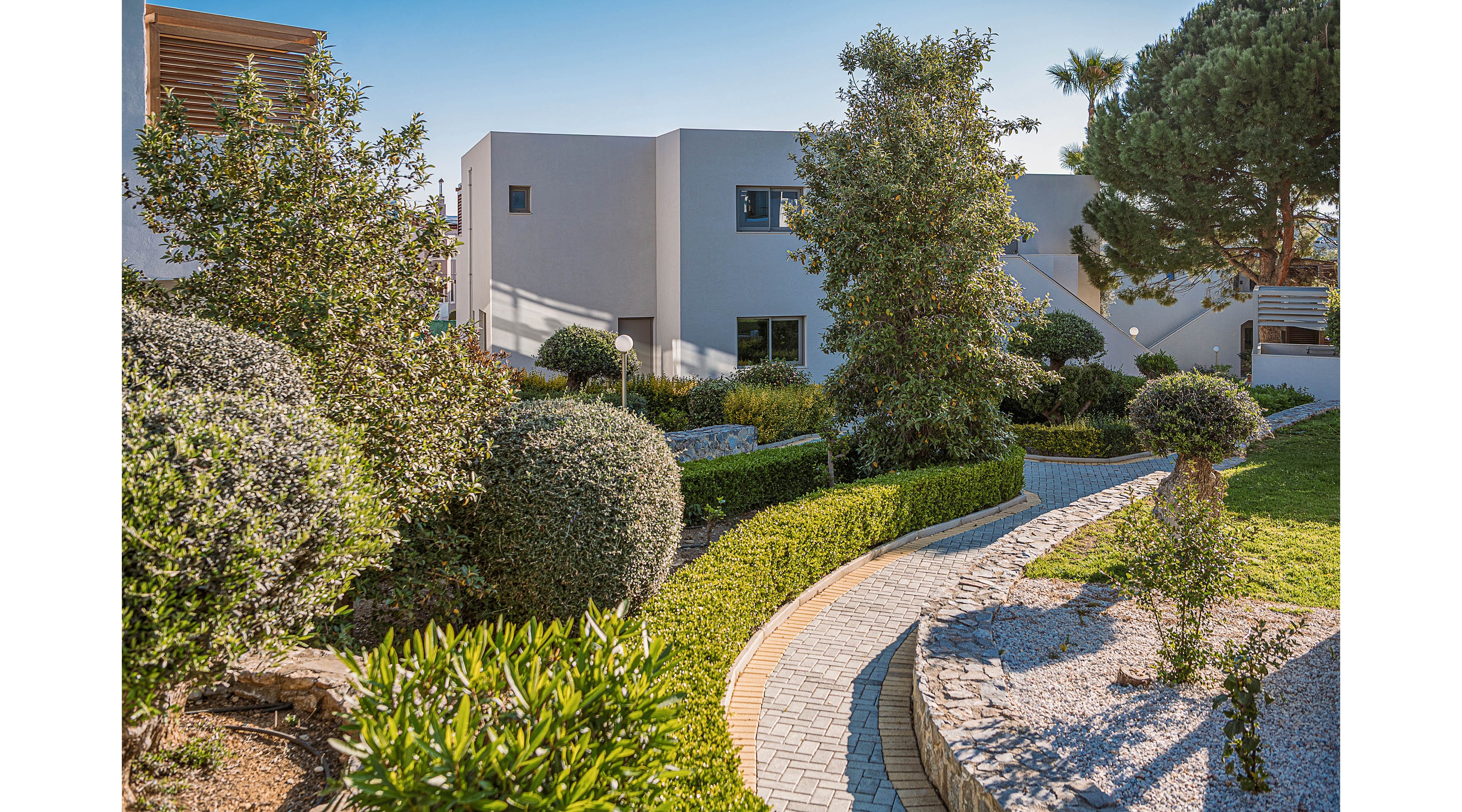 A modern white house is surrounded by a lush, well-maintained garden. A curved stone pathway leads through trimmed hedges, shrubs, and small trees toward the residence. Sunlight highlights the greenery and casts soft shadows, creating a warm and inviting atmosphere. The overall scene emphasizes contemporary architecture and carefully designed outdoor landscaping.