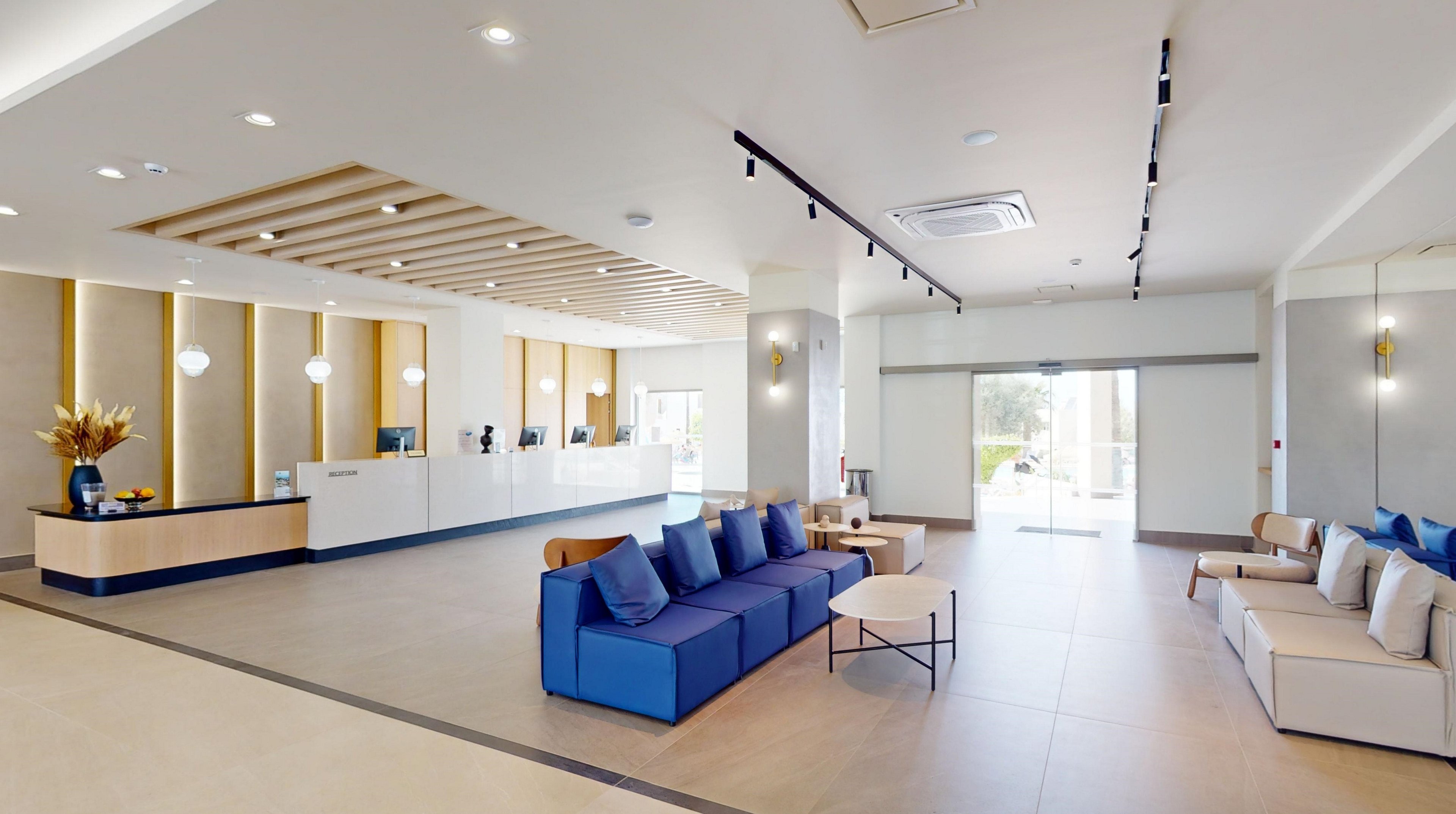 Spacious contemporary lobby interior featuring a long reception desk and multiple check-in stations. Comfortable sofas with blue and neutral cushions create a welcoming seating area in the foreground. Large floor-to-ceiling windows and glass doors allow natural light to fill the space. Clean lines, light wood accents, and recessed lighting emphasize a minimalist, modern design.