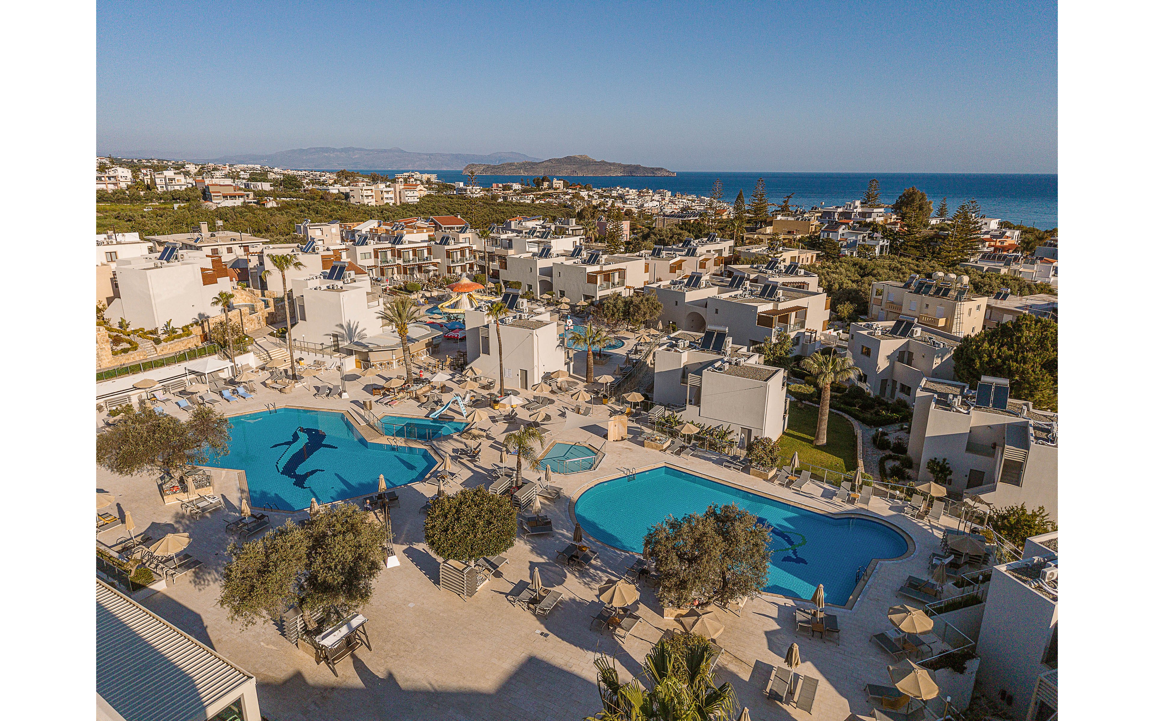 A large seaside resort complex is captured from above, showing multiple swimming pools surrounded by sun loungers and umbrellas. Modern low-rise buildings with flat roofs are arranged around landscaped paths and palm trees. The scene overlooks a coastal town stretching toward the blue sea and distant islands under a clear sky. The overall atmosphere is relaxed and summery, suggesting a popular vacation destination.