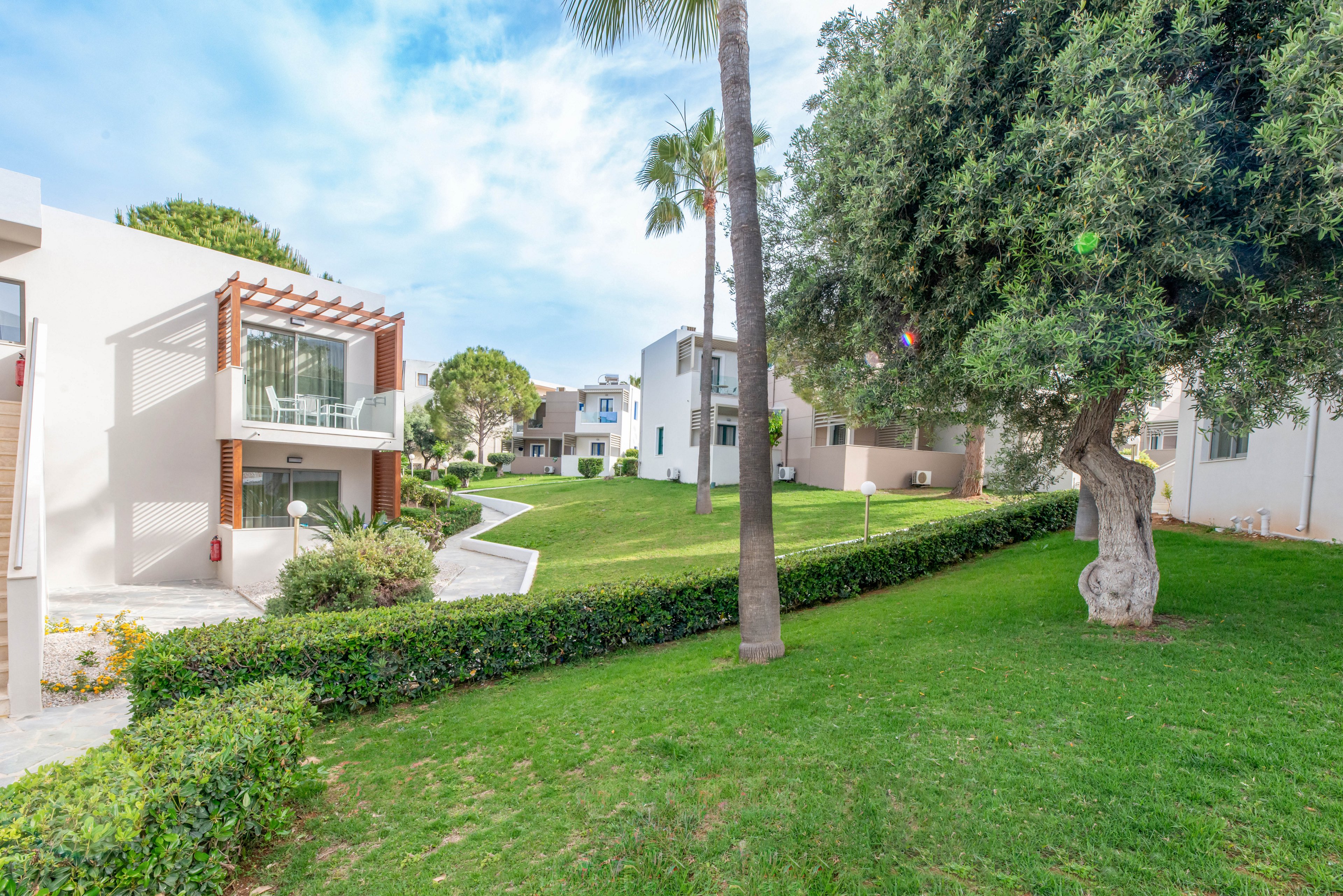 Contemporary white apartment buildings surround a spacious green courtyard. A well-maintained lawn, hedges, and mature trees create a peaceful residential or resort setting. Balconies and large windows suggest bright, airy interiors overlooking the shared garden. The scene is lit by soft daylight under a partly cloudy sky, emphasizing the relaxed, vacation-like atmosphere.