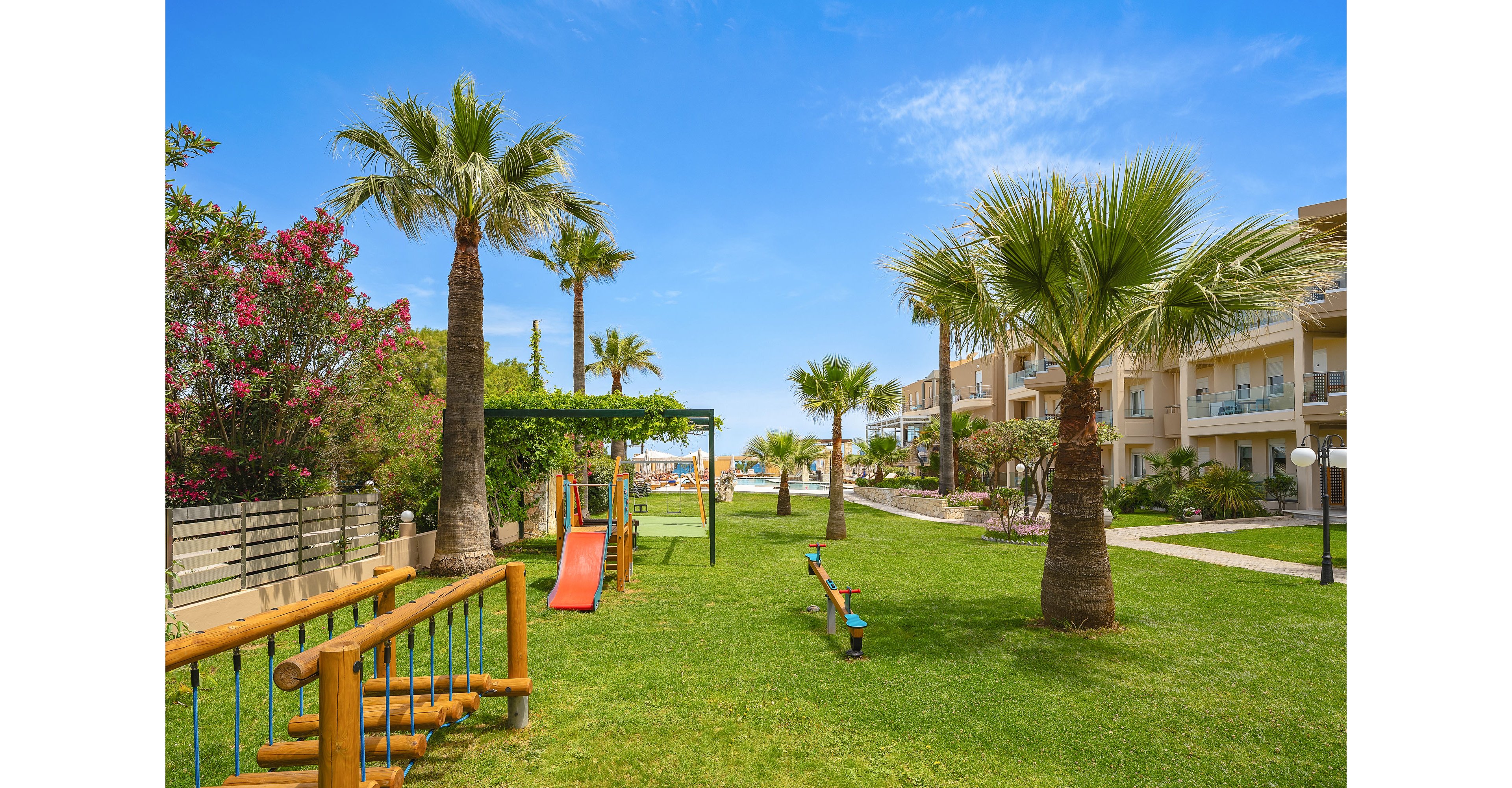 A bright, well-kept garden at a resort features a small children’s playground with slides and wooden climbing structures. Palm trees and flowering shrubs line the grassy path, creating a lush, tropical atmosphere. Apartment-style buildings with balconies are visible on the right, while outdoor lights and pathways guide guests through the area. The clear blue sky and distant view toward the sea suggest a warm, relaxing holiday setting.