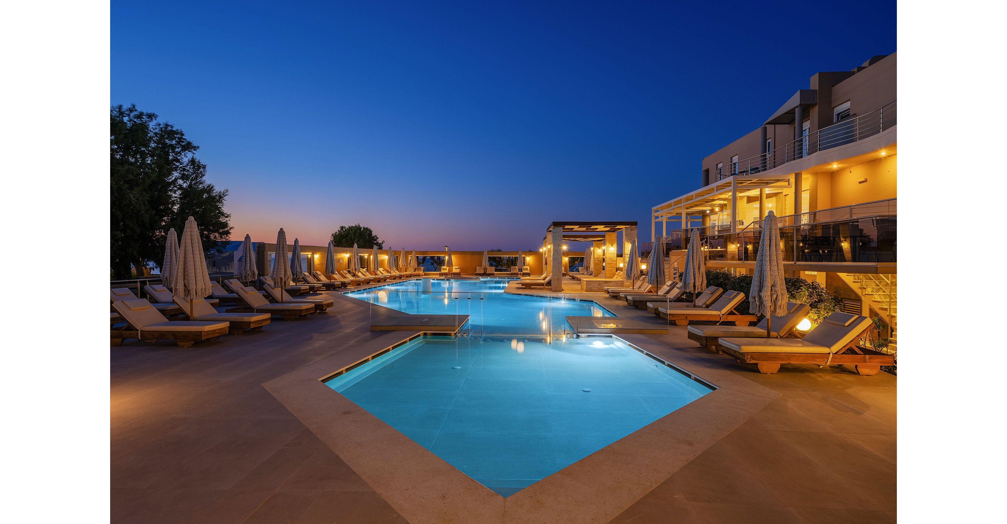 A modern resort swimming pool is illuminated at twilight, surrounded by sun loungers and umbrellas on a spacious terrace. The scene overlooks a calm horizon with the last light of sunset fading into a deep blue sky. Warm lights from the adjacent hotel building reflect on the water, creating a tranquil and inviting atmosphere. The clean architectural lines and soft lighting emphasize relaxation and upscale vacation living.