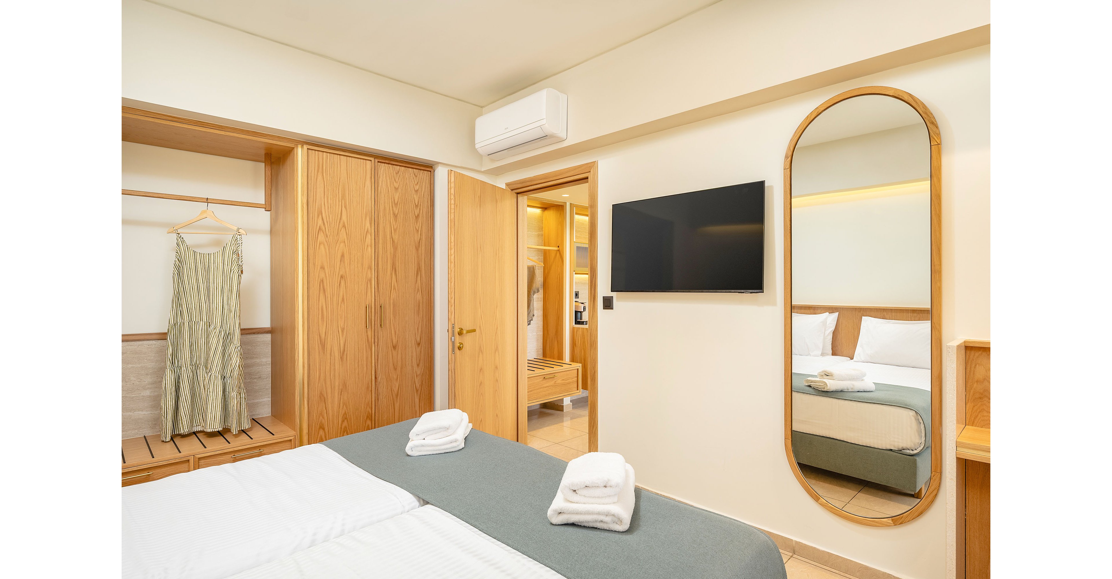Bright contemporary hotel room featuring a neatly made double bed with folded white towels on a gray bed runner. A large wooden wardrobe and open hanging space with a long dress create a warm, natural look. A wall-mounted flat-screen TV and tall oval mirror reflect the bed and bedside area, adding depth to the space. Soft neutral colors and clean lines give the room a calm, minimalist atmosphere.