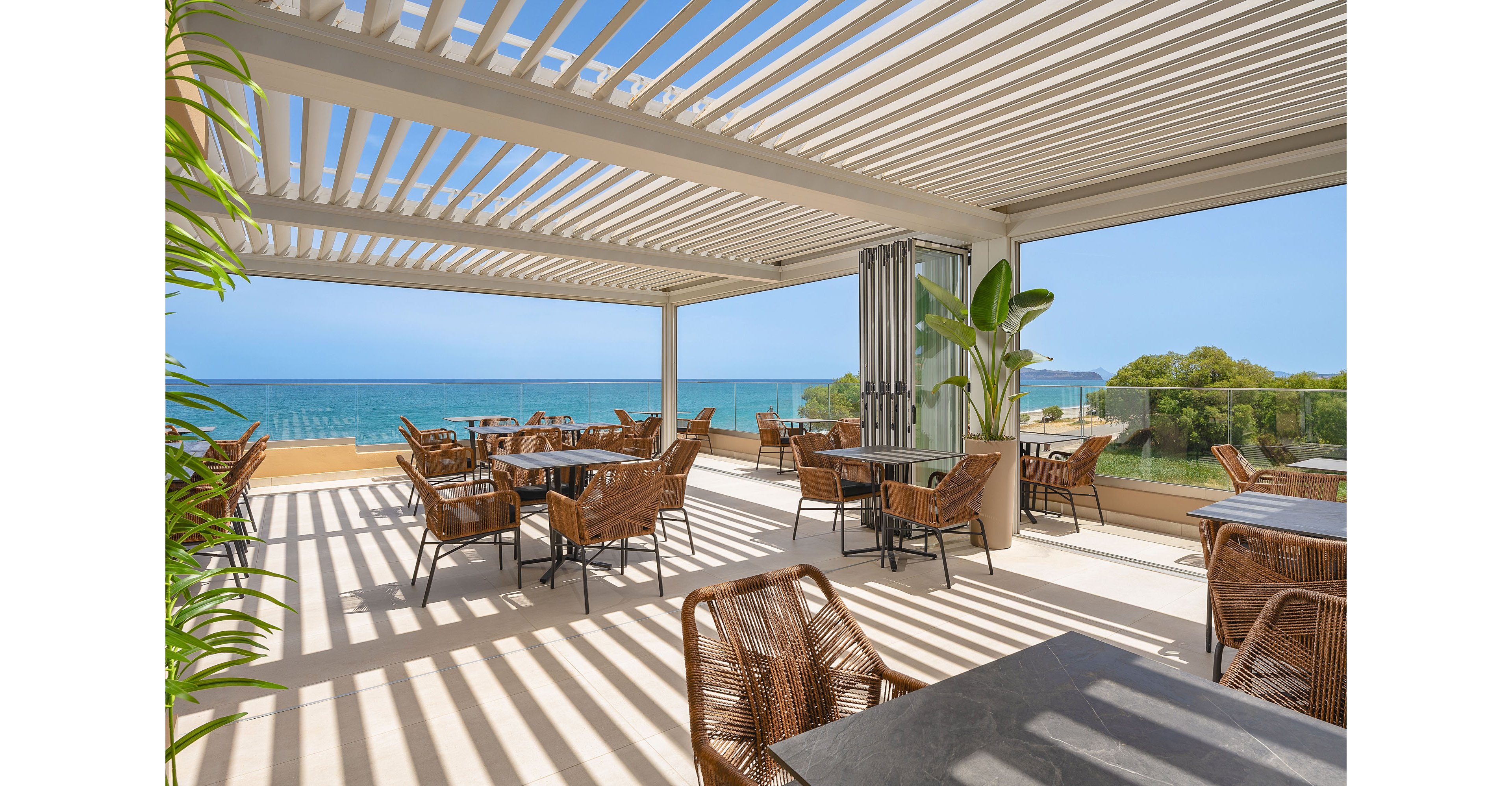 Open-air terrace café overlooks a bright blue ocean under a clear sky. Contemporary pergola slats cast strong linear shadows across the floor and tables. Wicker chairs surround dark stone tables, creating a relaxed yet stylish dining area. Green plants add a touch of nature to the clean, modern architecture and coastal setting.