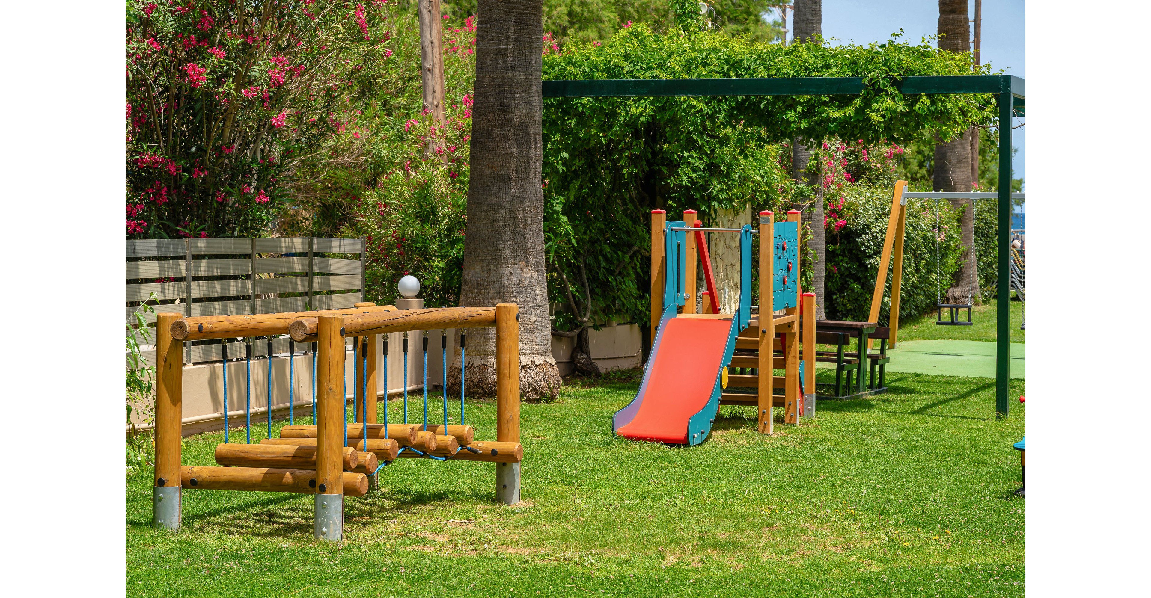 A small outdoor playground features wooden climbing equipment and a bright red and blue slide on a green lawn. The play area is surrounded by palm trees, flowering bushes, and a fence, creating a cozy garden setting. A shaded pergola structure with greenery covers part of the playground, and nearby chairs suggest a space for adults to sit. The scene is lit by clear daylight, giving the image a cheerful and inviting atmosphere.