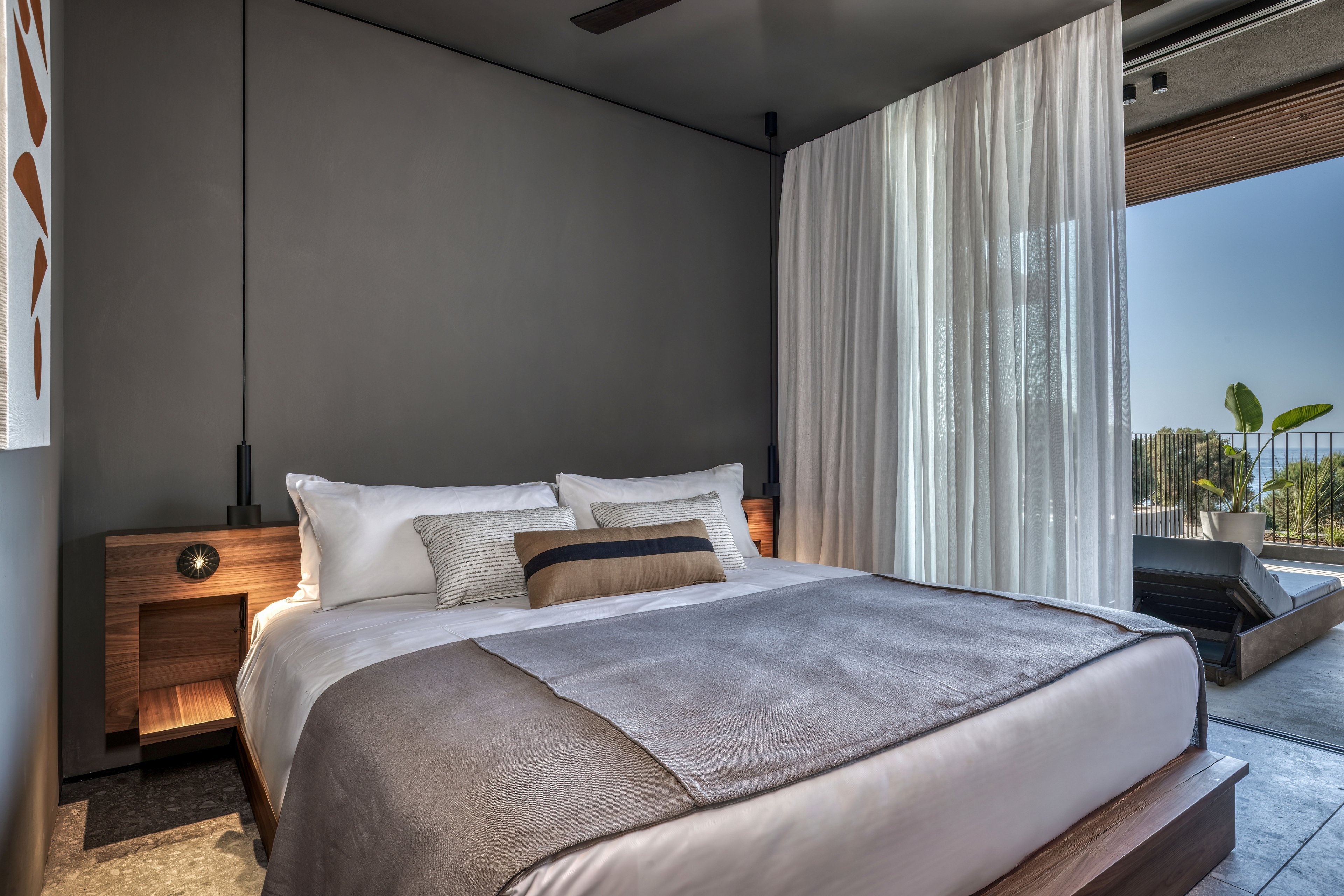 A contemporary bedroom features a large neatly made bed with white linens and neutral-toned pillows and throw. Wooden nightstands with built-in lights flank the bed against a smooth gray wall. Sheer white curtains partially open to reveal an outdoor terrace with seating and greenery. Natural light streams in from the balcony, enhancing the calm, modern atmosphere of the space.