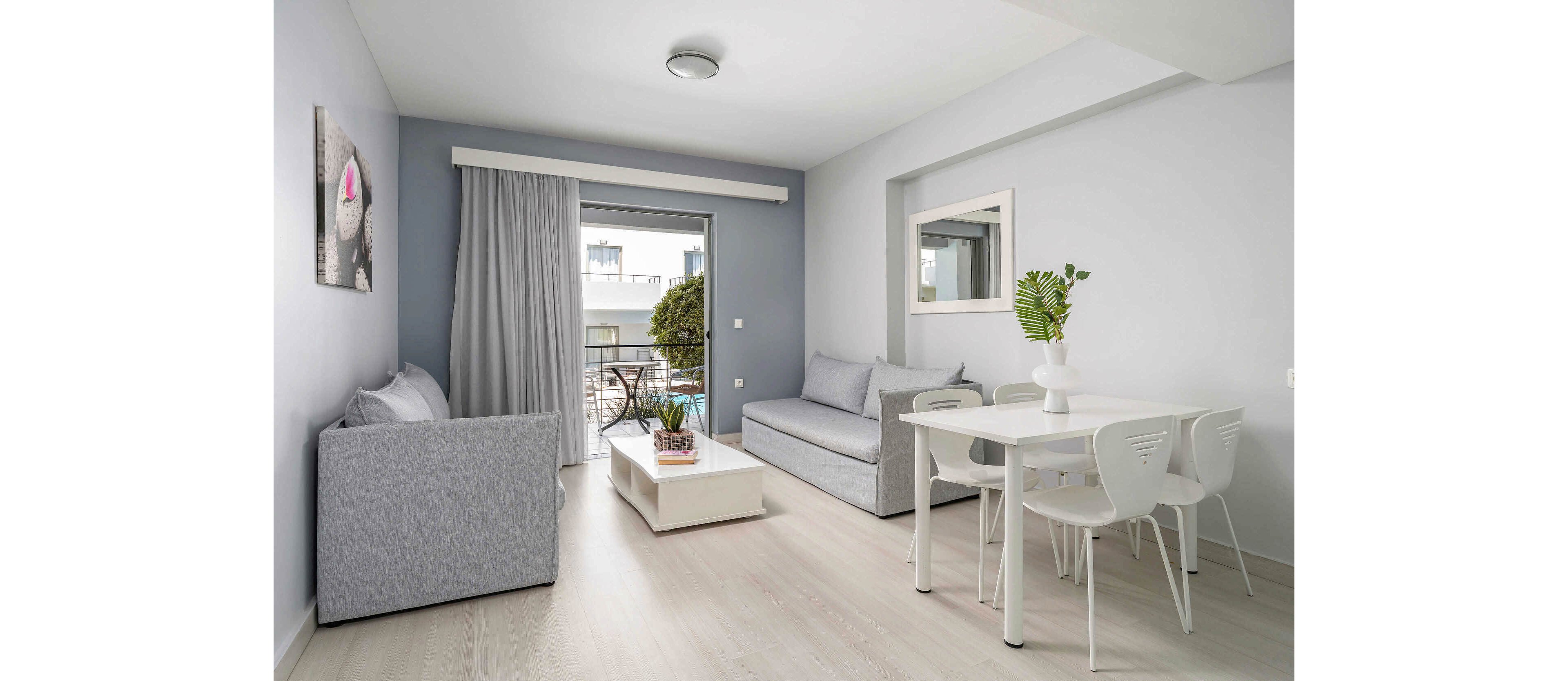 Contemporary living and dining area with minimalist furniture in soft grey and white tones. The room features two sofas, a white dining table with four chairs, and a low coffee table with a plant centerpiece. Large sliding glass doors open to a small balcony with outdoor seating and greenery. Light wood flooring and pale blue walls create an airy, relaxing atmosphere.
