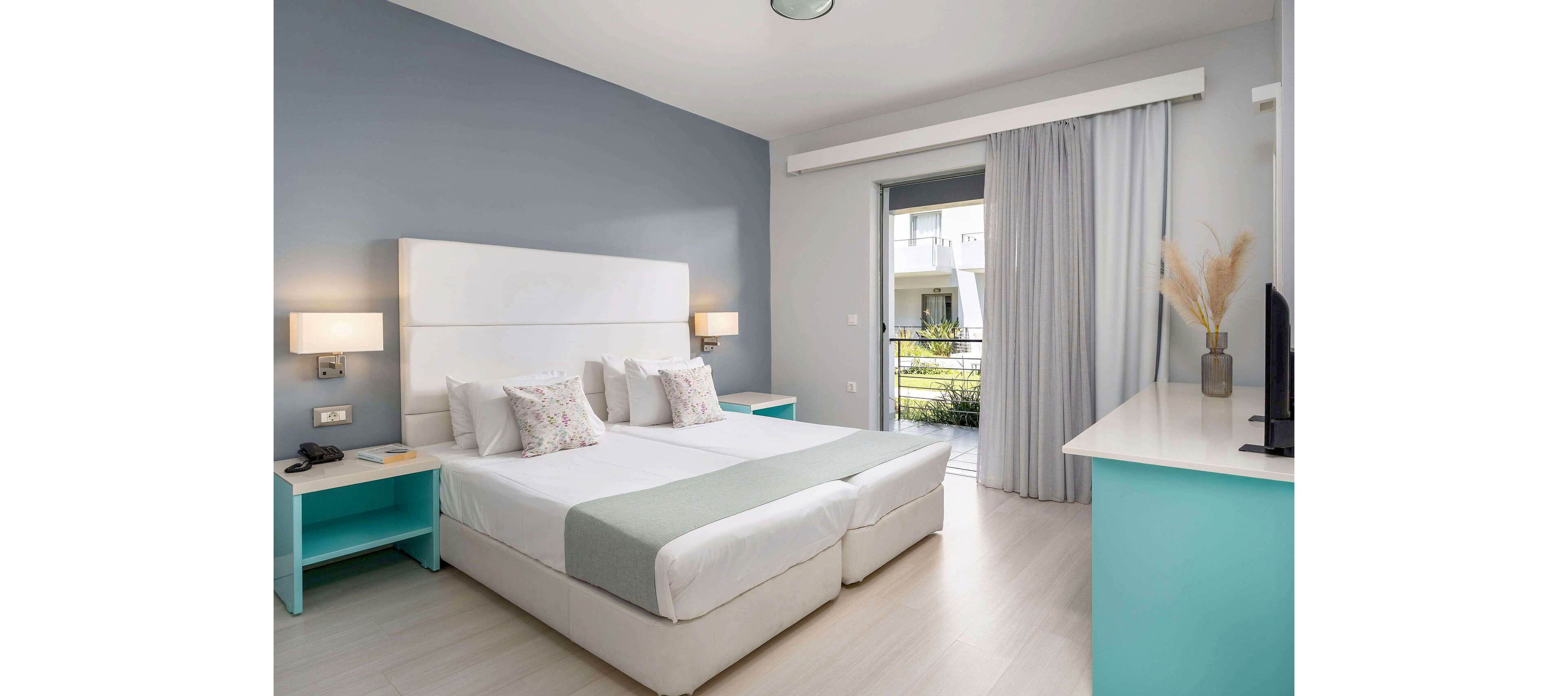 Contemporary hotel room featuring two single beds pushed together with a padded white headboard. The space is decorated in soft pastel tones with turquoise bedside tables and a matching desk. Large glass doors with sheer curtains open to a balcony overlooking a green courtyard. Minimal decor, including floral pillows and a vase with dried grasses, enhances the clean and relaxing atmosphere.