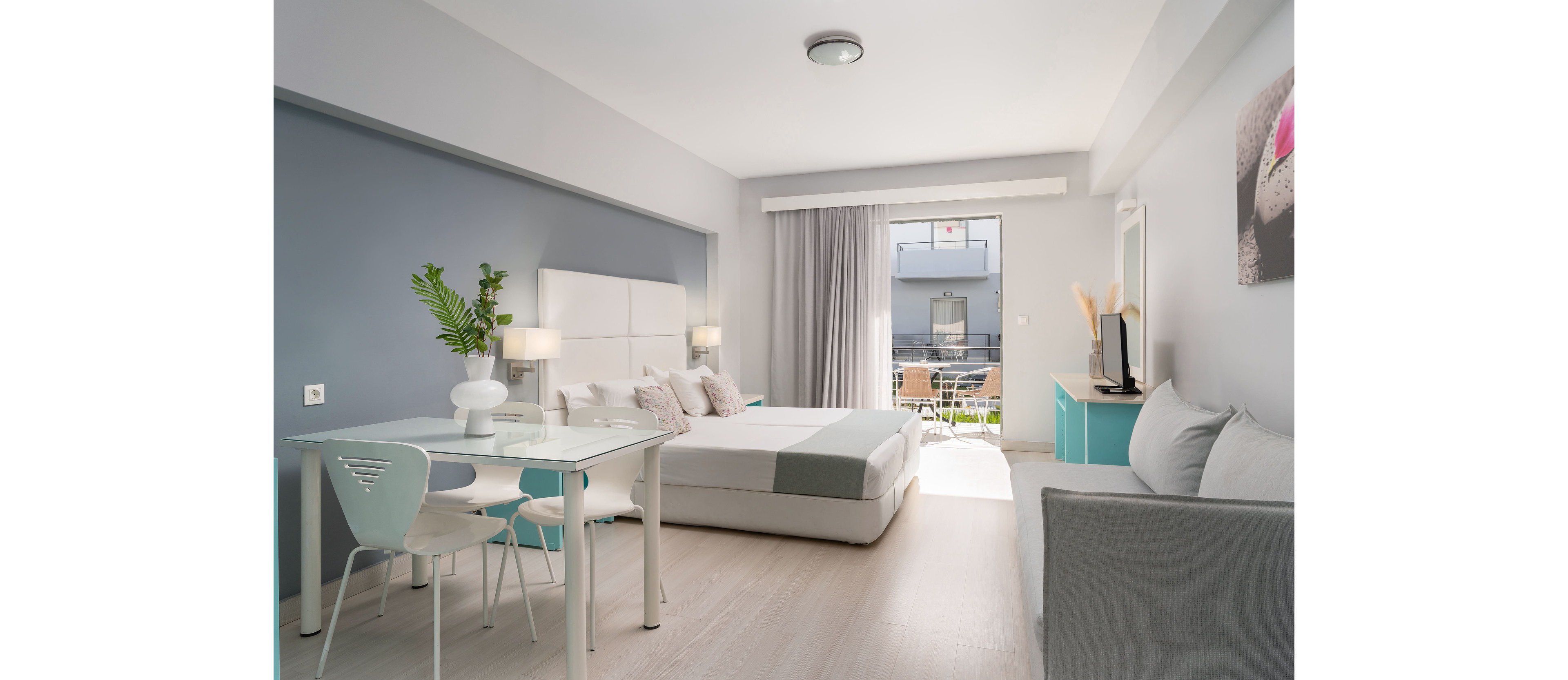A spacious, modern hotel room features a neatly made double bed with light-colored linens and accent pillows. The interior includes a small dining table with chairs, a sofa, and a TV on a turquoise cabinet. Soft natural light enters through large glass doors that open to a balcony overlooking a courtyard. The decor uses a calm palette of white, gray, and pastel blue with minimal, contemporary furnishings.