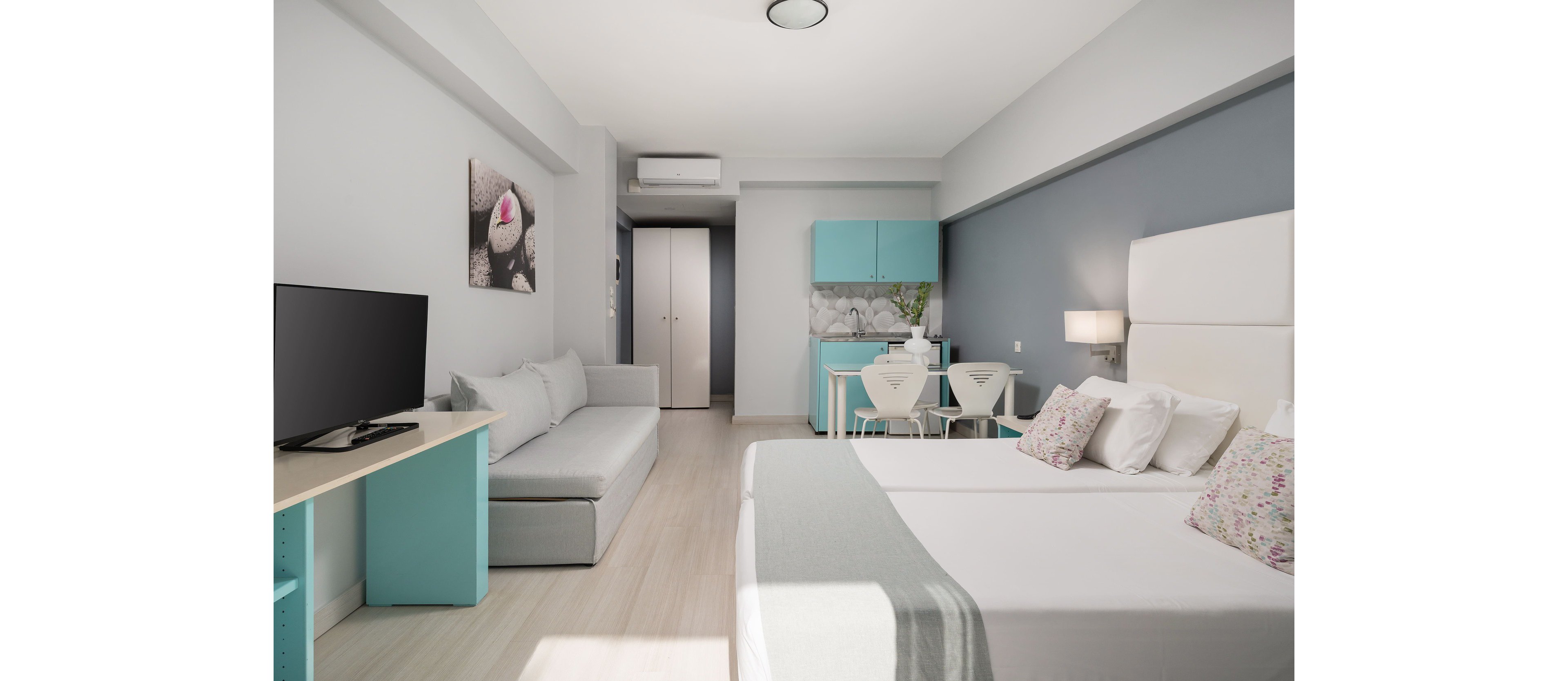 A bright modern studio apartment features a neatly made double bed with decorative pillows in the foreground. To the left, a light gray sofa faces a flat-screen television on a minimalist stand. At the back of the room, a compact kitchenette with turquoise cabinets and a small dining table with white chairs adds functionality. Soft neutral walls with a blue accent stripe and pastel accents create a calm, contemporary atmosphere. The space appears clean, organized, and designed for comfortable short-term stays.
