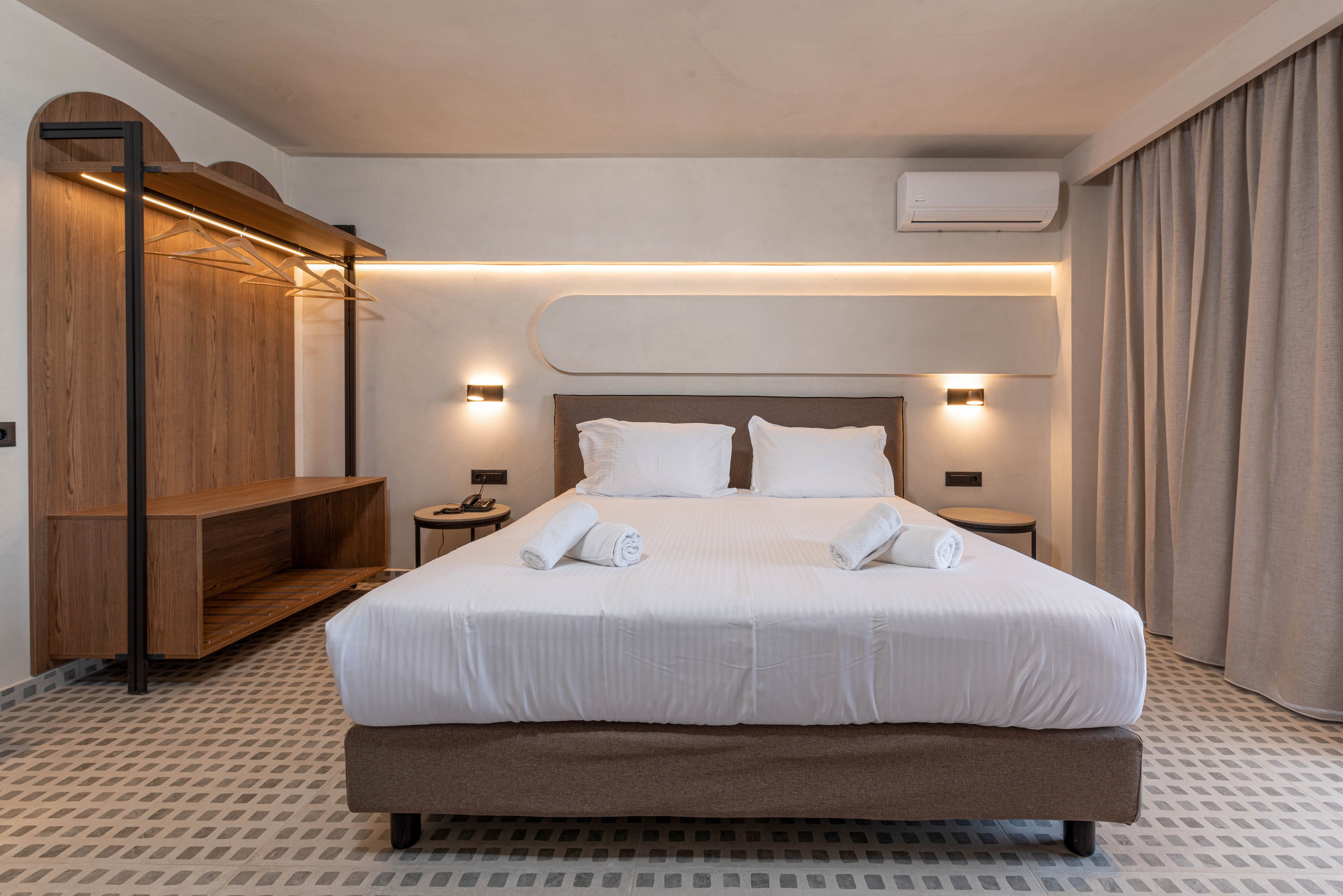Contemporary hotel room featuring a double bed with crisp white linens and neatly rolled towels placed on top. The space includes a minimalist open wardrobe and shelving unit in wood on the left side. Soft ambient lighting runs along the wall above the bed, complemented by two wall-mounted bedside lamps. Neutral tones, patterned flooring, and floor-to-ceiling curtains create a calm, inviting atmosphere.