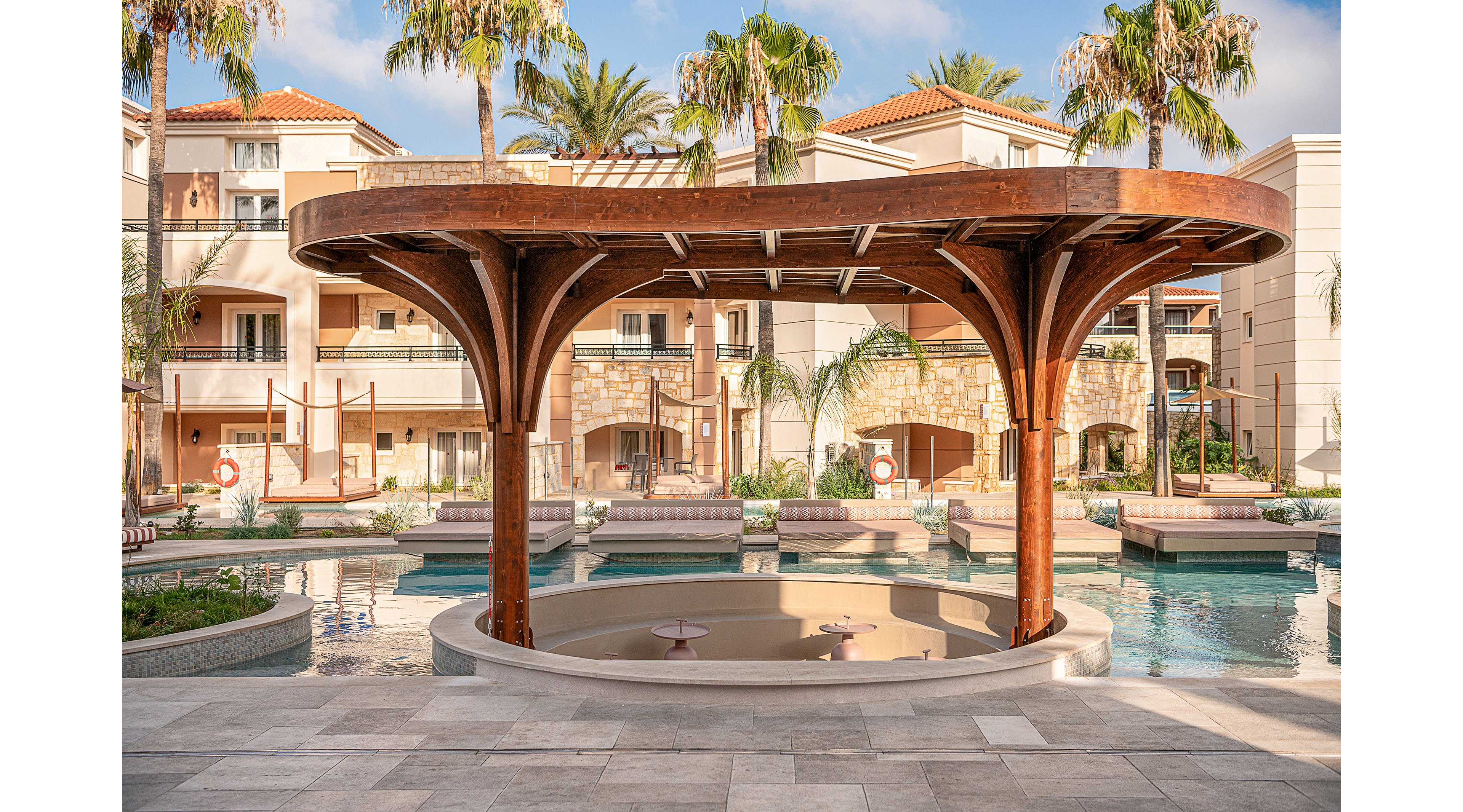 An open wooden pavilion stands at the center of a resort-style swimming pool area. Surrounding it are lounge seating, palm trees, and multi-story residential or hotel buildings with balconies. The scene is bright and sunny, with clear reflections on the calm water and a relaxed vacation atmosphere. Architectural details include stone accents, tiled roofs, and neatly arranged walkways around the pool.