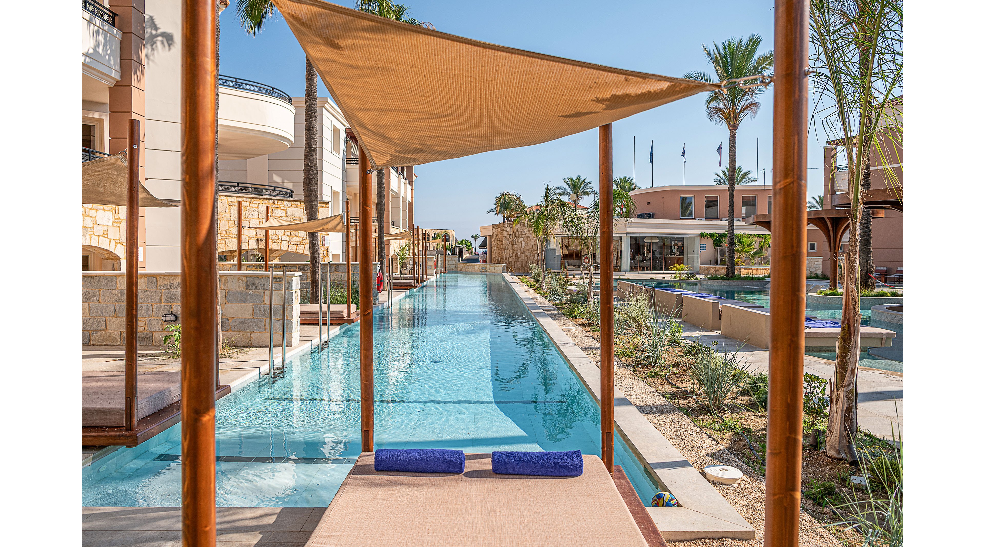 A long, narrow swimming pool runs between modern resort buildings, framed by wooden posts and a fabric canopy walkway. Two blue rolled towels rest on a sunbed at the edge of the pool, suggesting a relaxing vacation setting. Palm trees, desert-style landscaping, and clear blue water create a bright, tranquil atmosphere. In the background, flags and additional structures indicate a large, upscale hotel complex under sunny skies.