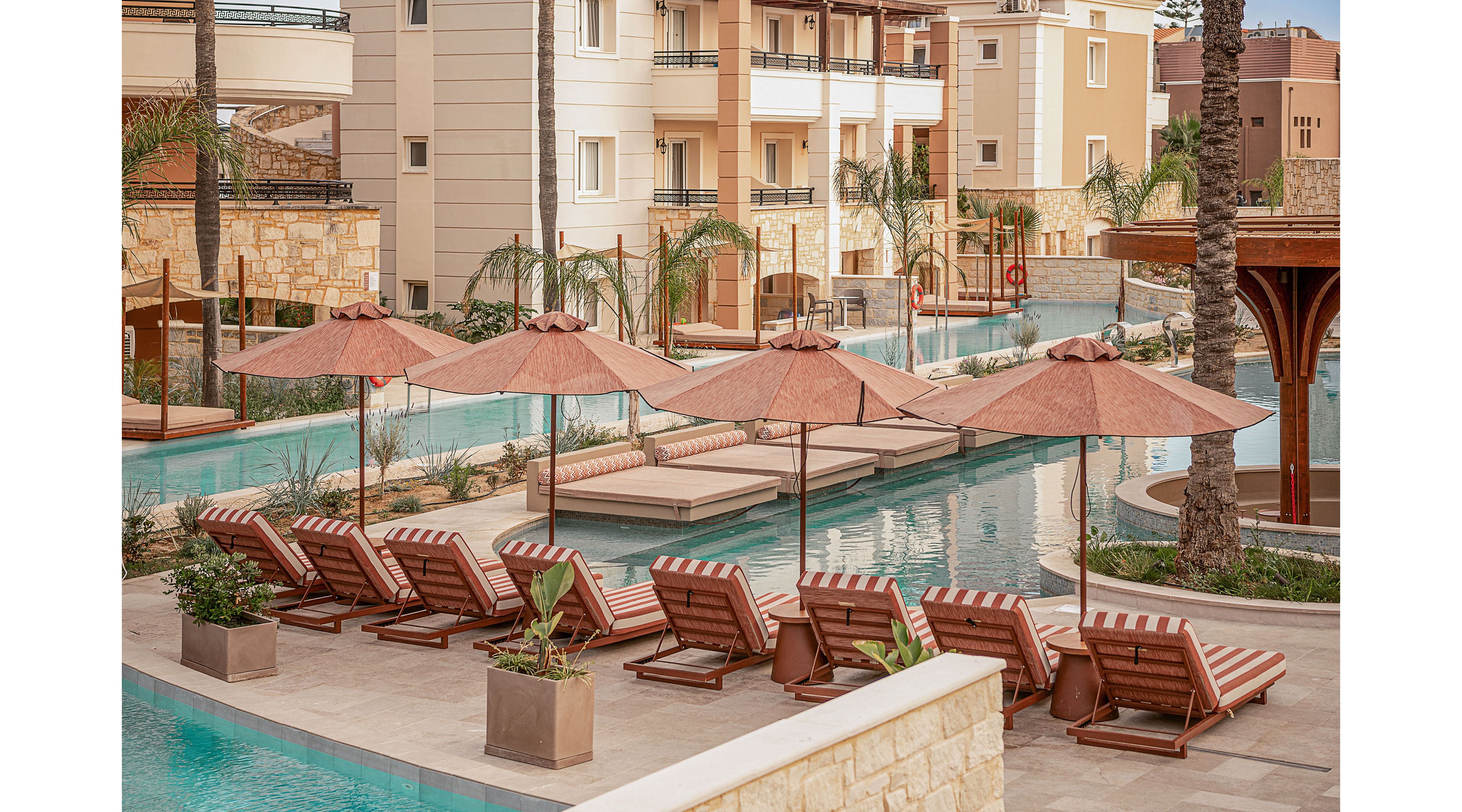 A row of cushioned sun loungers with red and white stripes lines the edge of a resort swimming pool. Matching umbrellas provide shade along the curved pool, which features bridges and platforms over the water. Palm trees and stone planters add a tropical touch to the scene. In the background, multi-story residential or hotel buildings frame the tranquil outdoor area.
