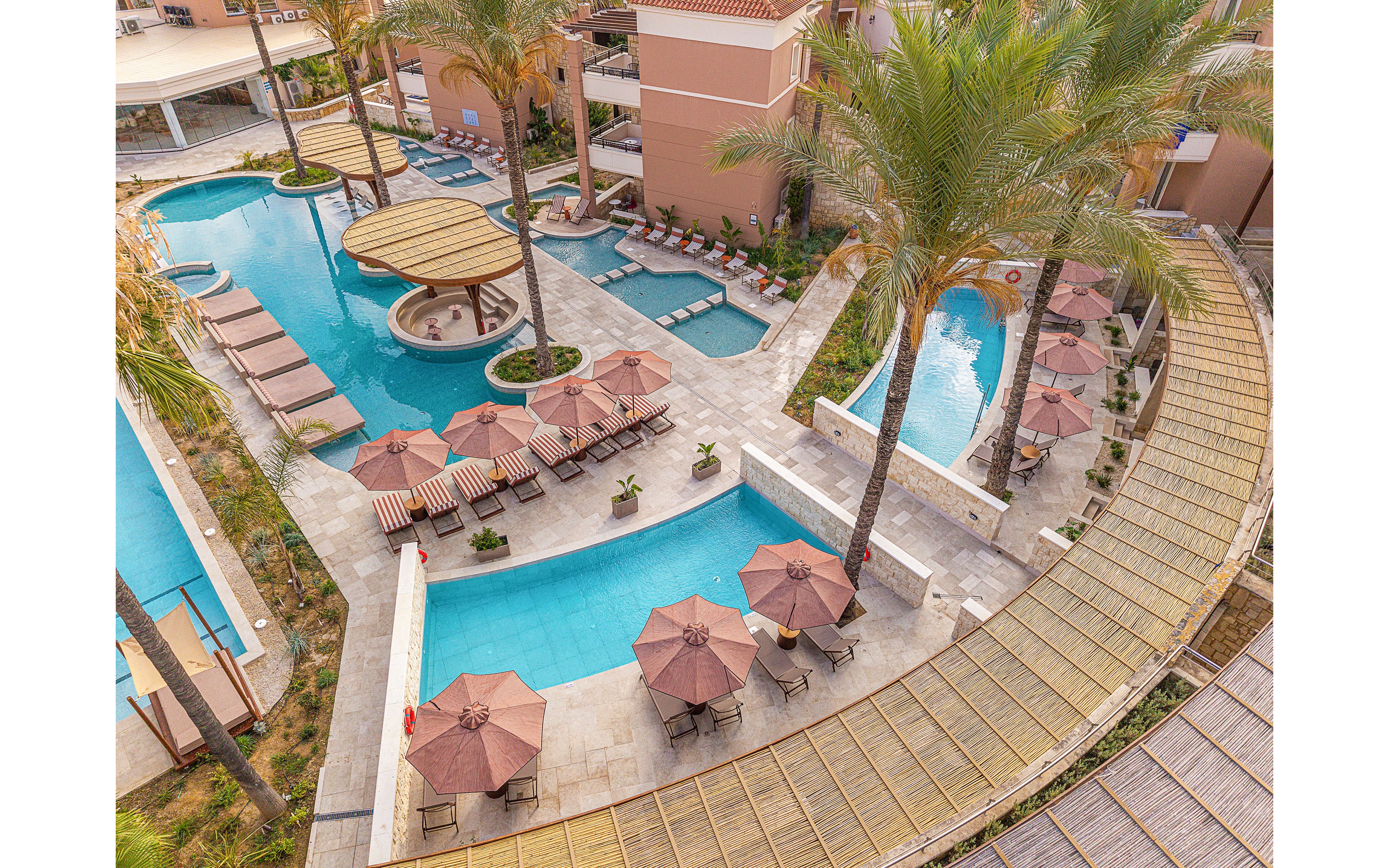 A large resort-style swimming pool complex is seen from above, featuring multiple interconnected pools and sunbathing areas. Lounge chairs and umbrellas line the poolside terraces, with several shaded seating zones arranged around the water. Tall palm trees and landscaped walkways add a tropical atmosphere to the scene. The surrounding buildings and curved pathways frame the leisure area, emphasizing a relaxing vacation setting.