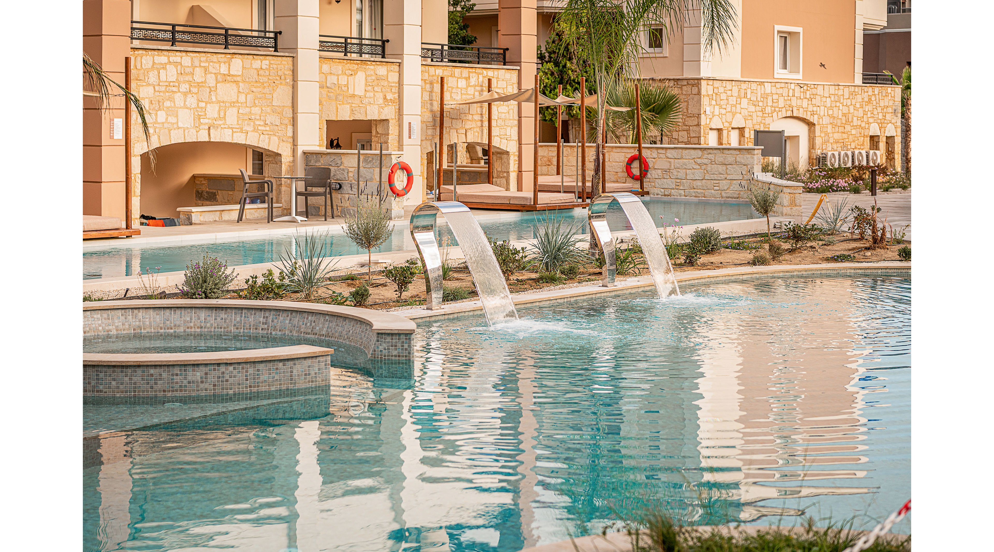 An inviting resort-style swimming pool is shown in front of a modern building with stone and stucco walls. Curved metal fountains pour water into the clear blue pool, creating gentle ripples on the surface. Palm trees, shrubs, and neatly arranged landscaping line the poolside, along with safety rings mounted on posts. The overall scene suggests a relaxing vacation atmosphere in a warm, sunny climate.