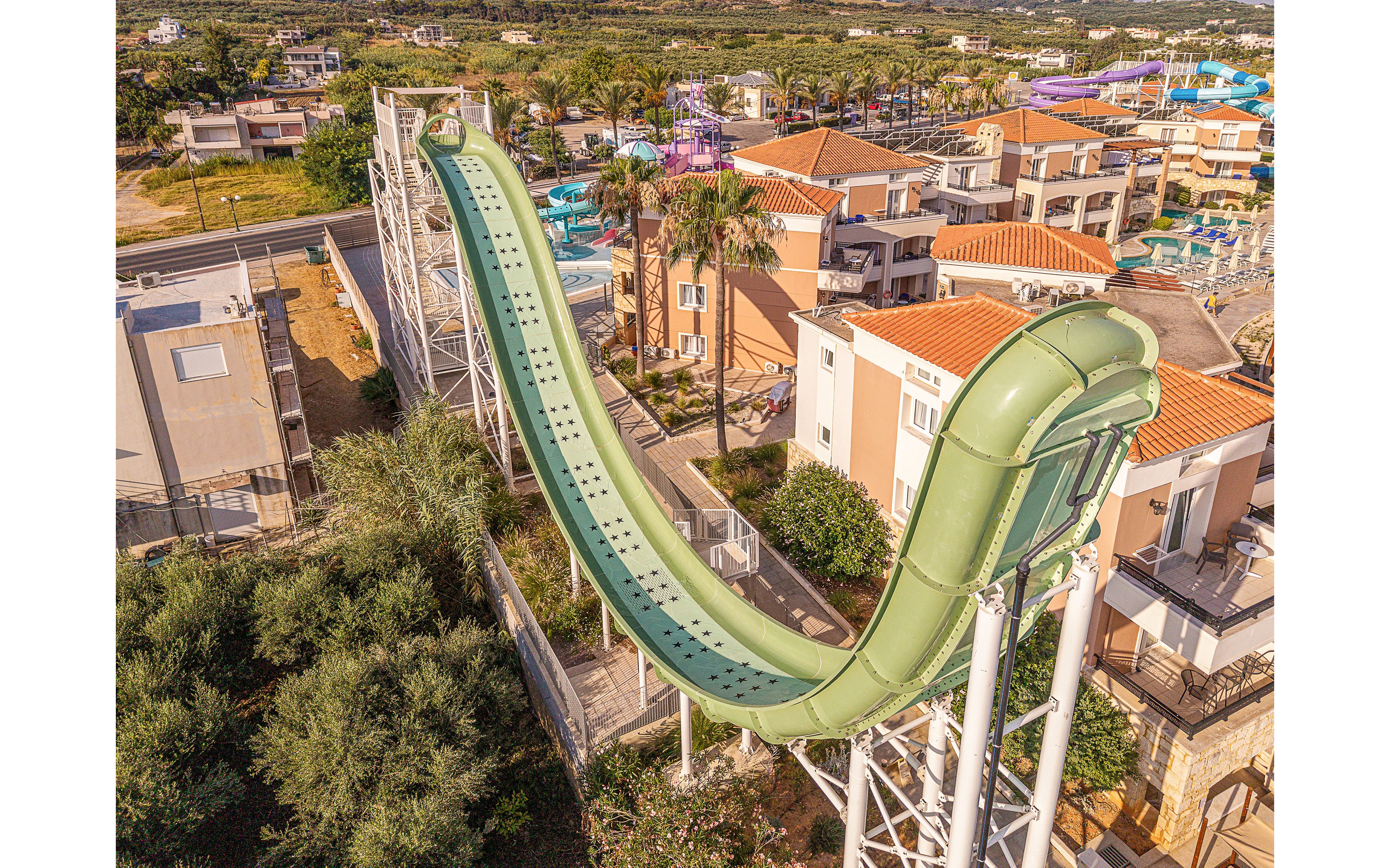 A large green water slide towers above a resort complex with multiple buildings and palm trees. The slide curves steeply downward and upward, supported by a white metal structure. Surrounding the slide are residential-style accommodations with orange-tiled roofs and nearby swimming pools. The scene is captured from an elevated viewpoint, highlighting the scale of the attraction within the sunny vacation setting.