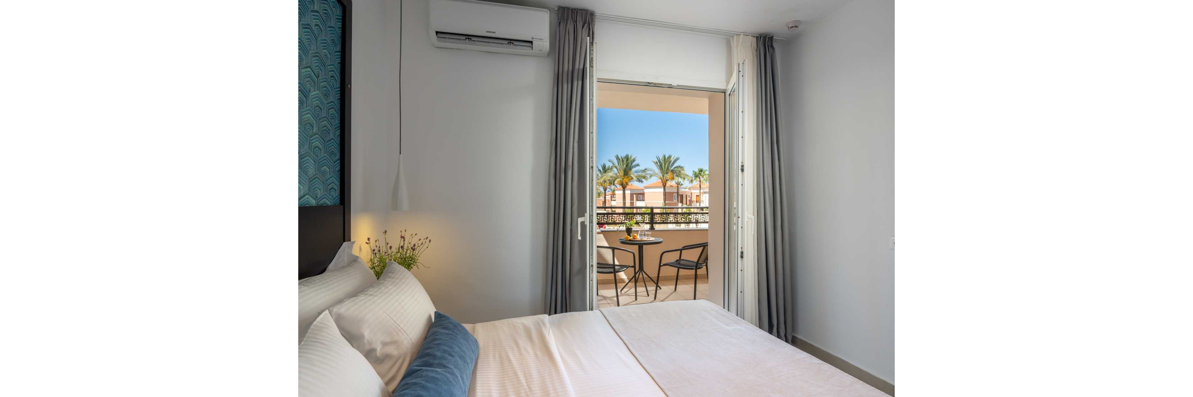 A modern hotel bedroom features a neatly made bed with white linens and accent pillows. Floor-to-ceiling curtains frame an open doorway leading to a sunny balcony. Outside, a small table with chairs overlooks palm trees and resort buildings under a clear blue sky. The scene conveys a relaxed vacation atmosphere with clean, contemporary decor.