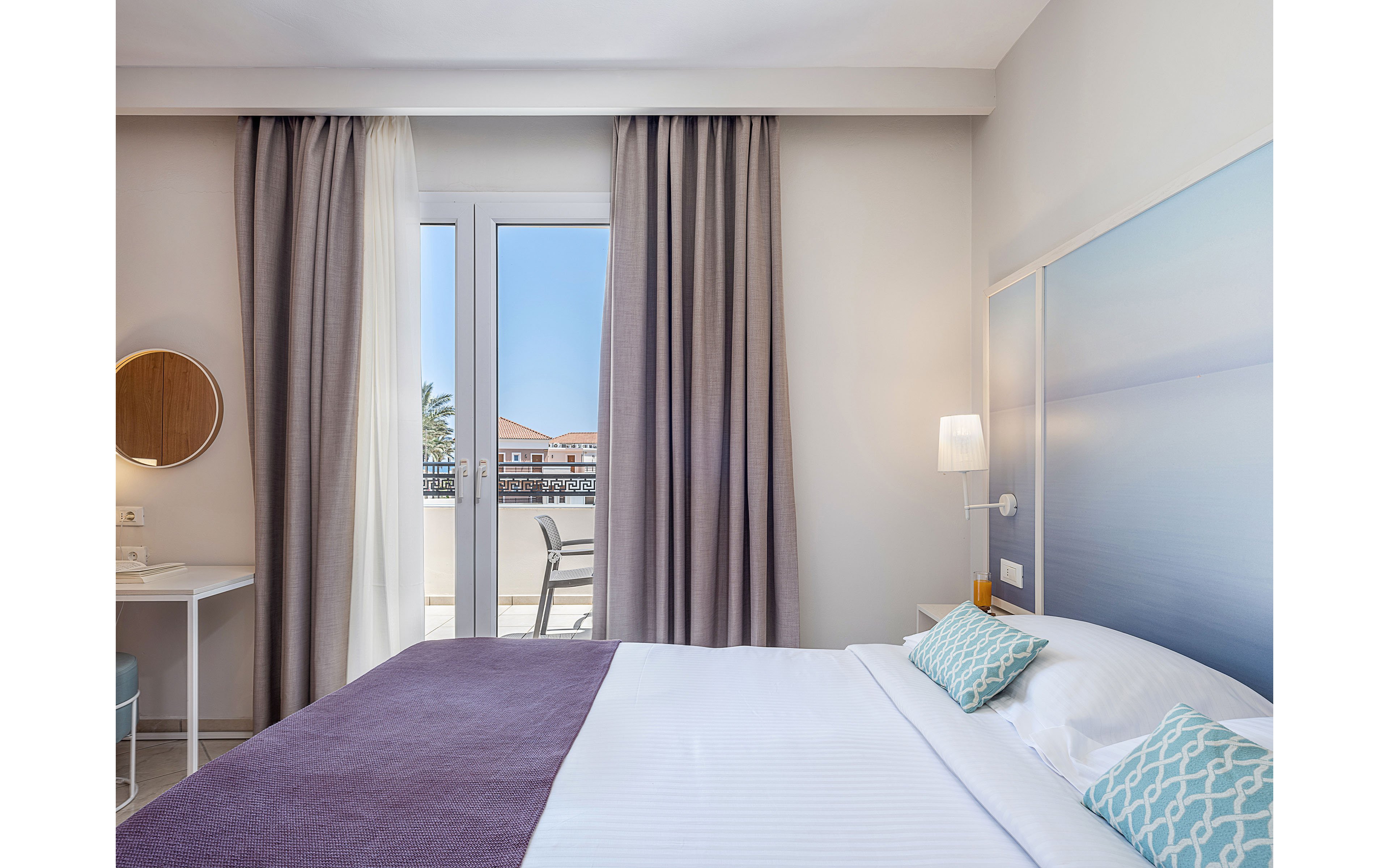 Contemporary hotel bedroom with a neatly made double bed faces a balcony with large glass doors. Soft gray and beige curtains frame the bright outdoor view of buildings and blue sky. The room features a minimalist desk with round mirror, wall-mounted lamp, and a serene seascape artwork above the bed. Light colors, clean lines, and patterned accent pillows create a calm, inviting atmosphere.