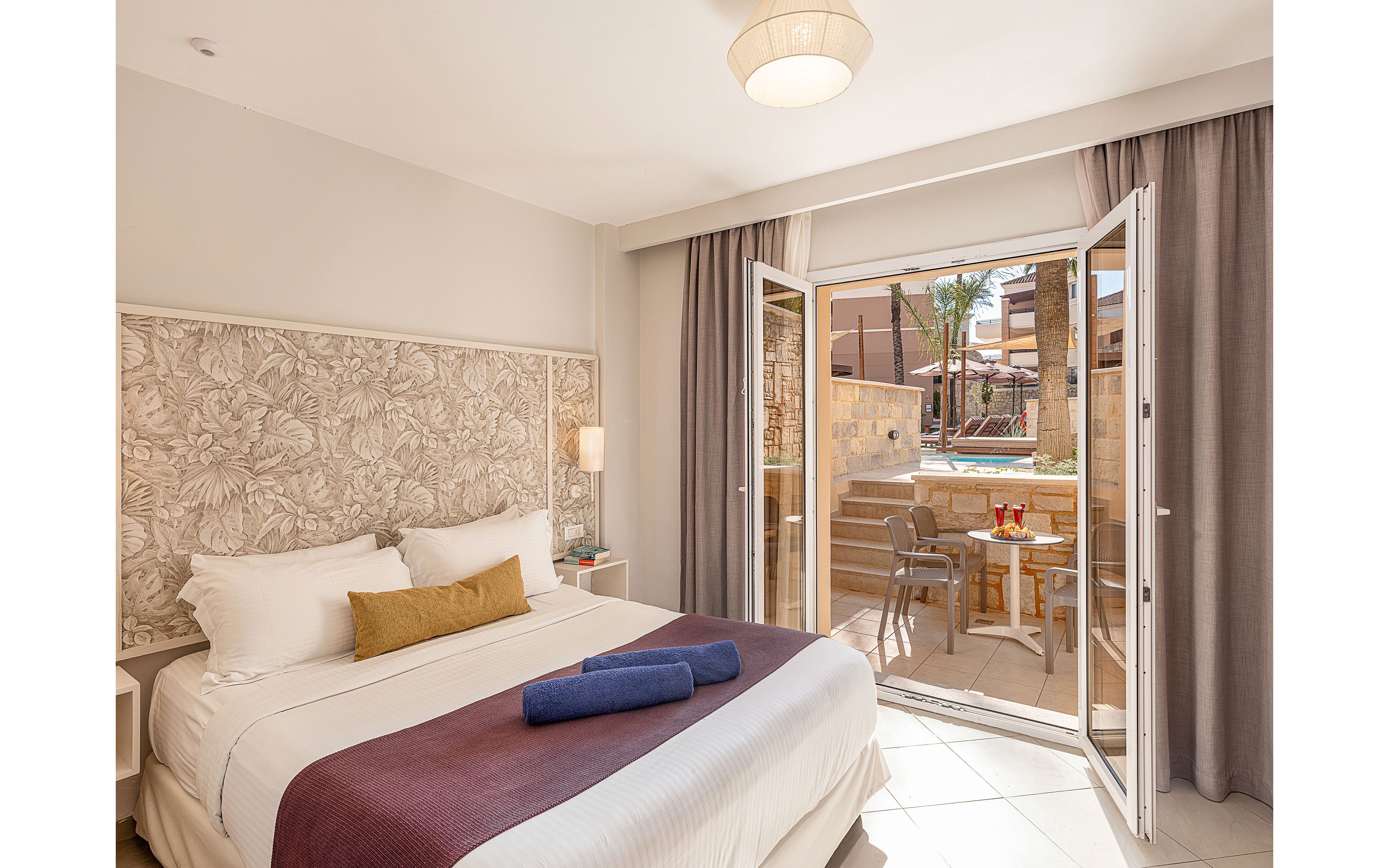 A sunlit hotel bedroom features a neatly made double bed with white linens, a mustard accent pillow, and a folded blue towel. Decorative floral wall paneling and soft neutral curtains add a modern, cozy feel to the space. Through the open glass doors, a private terrace is visible with a small table, chairs, and colorful drinks, overlooking a stone-walled pool area. The overall scene suggests a relaxing vacation stay in a warm, resort-like setting.