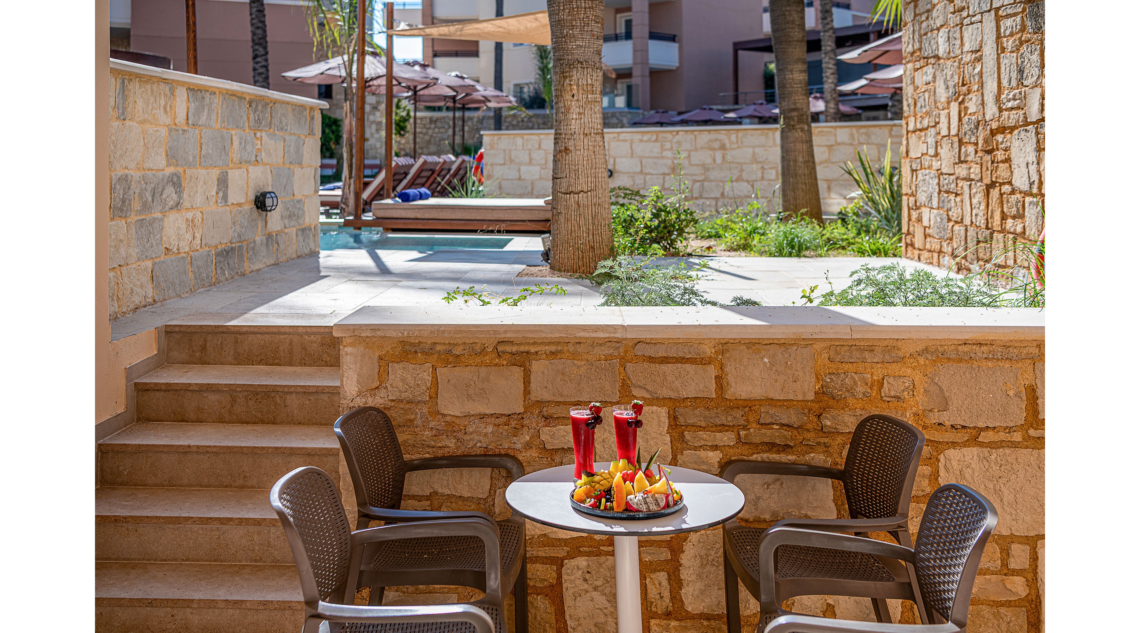 An outdoor resort patio features a small round table set with tall red drinks and a platter of fresh fruit. Four wicker-style chairs surround the table, inviting guests to sit and relax. Stone walls, steps, and tropical plants frame the scene, leading toward a pool area with sun loungers and umbrellas in the background. The bright daylight and warm colors create a summery, vacation atmosphere.