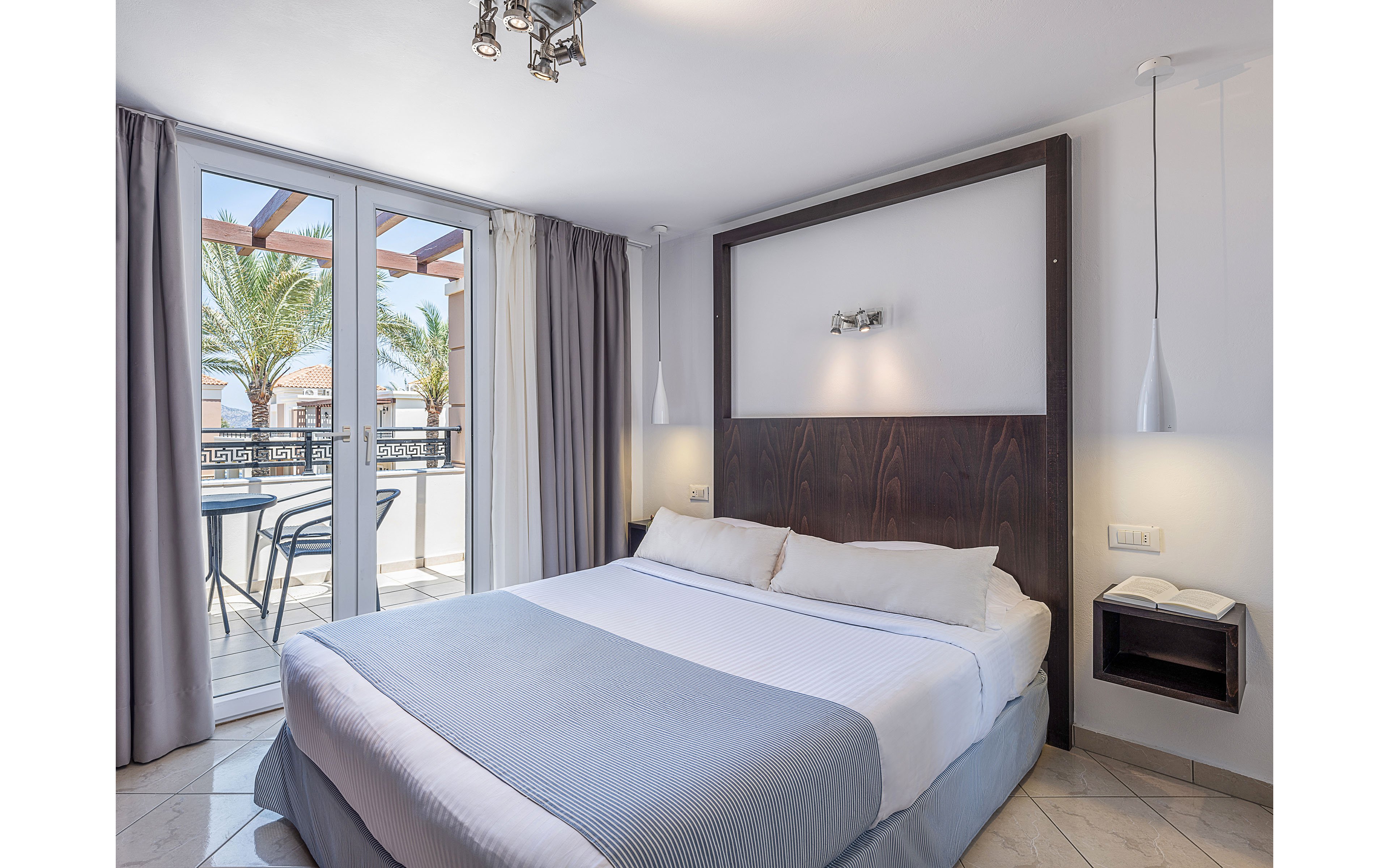 Modern hotel bedroom featuring a neatly made double bed with white linens and a light gray blanket. Large glass doors open to a balcony with outdoor chairs and a view of palm trees and nearby buildings. The room includes contemporary pendant lights, a dark wood headboard, and neutral curtains. Overall, the space feels airy, minimalist, and inviting for guests.