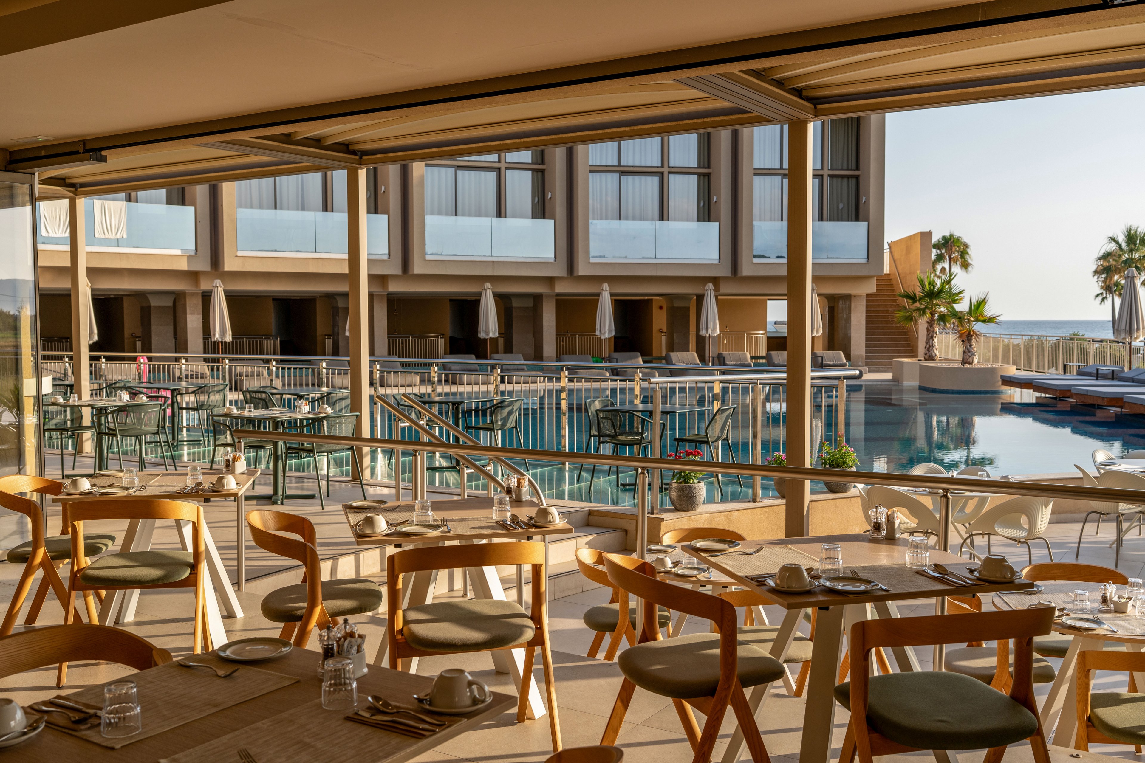 An open-air resort restaurant terrace features neatly arranged wooden chairs and tables set for dining. Beyond the terrace, a large swimming pool is surrounded by sun loungers, umbrellas, and modern hotel buildings. The scene is bathed in warm sunlight, with a clear view toward the sea and palm trees in the background. The overall atmosphere suggests a relaxed vacation setting ideal for outdoor meals and leisure.