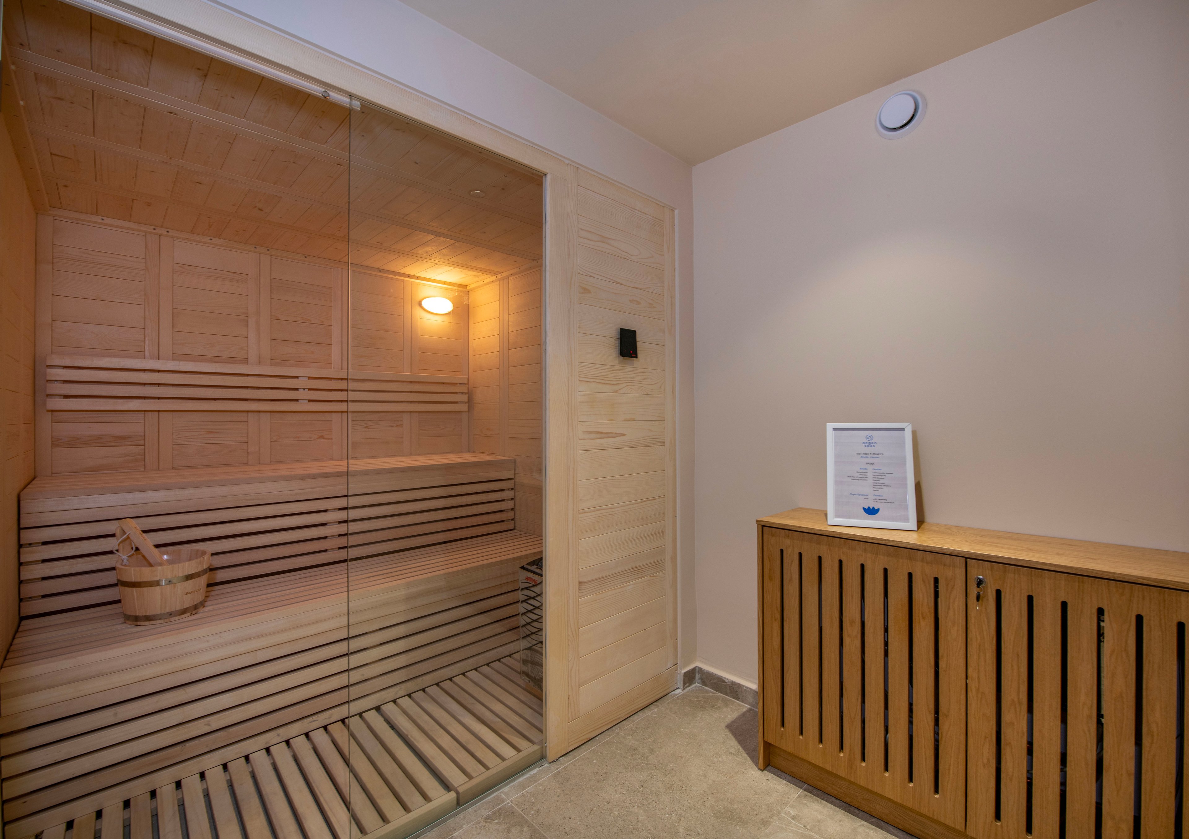 Image of a contemporary wooden sauna with tiered benches and a glass door. A wooden bucket sits on the bench inside the sauna, illuminated by a warm wall light. Outside the sauna, there is a wooden cabinet with vertical slats and a framed document on top. The space has neutral-colored walls and stone flooring, creating a clean and relaxing wellness environment.