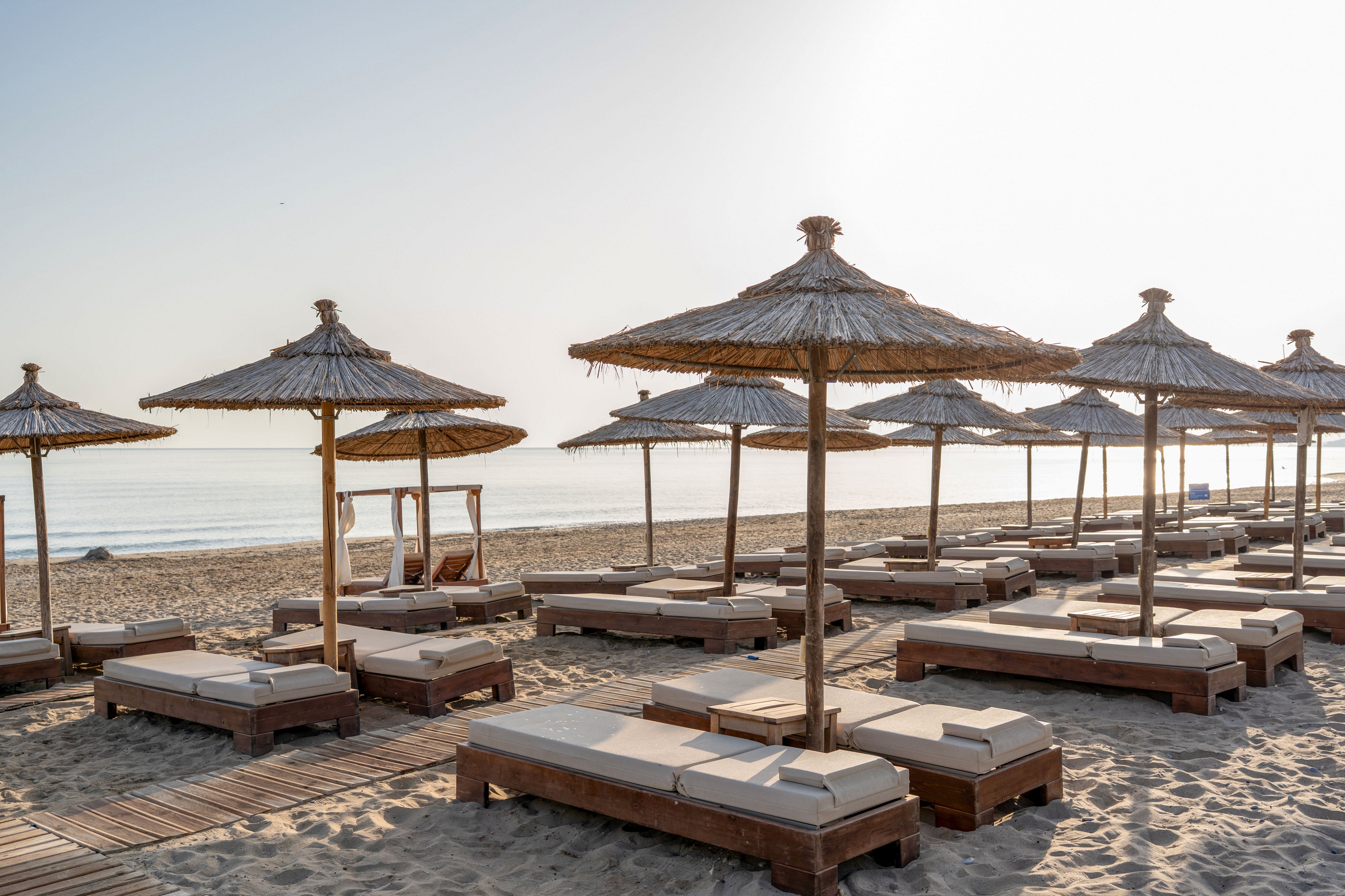 A tranquil beach resort features rows of cushioned sun loungers under straw parasols on sandy shore. Wooden walkways lead between the loungers toward the calm sea in the background. The water is smooth and reflective under the soft early morning or late afternoon light. The scene feels peaceful and inviting, with no people present and a clear, bright sky.