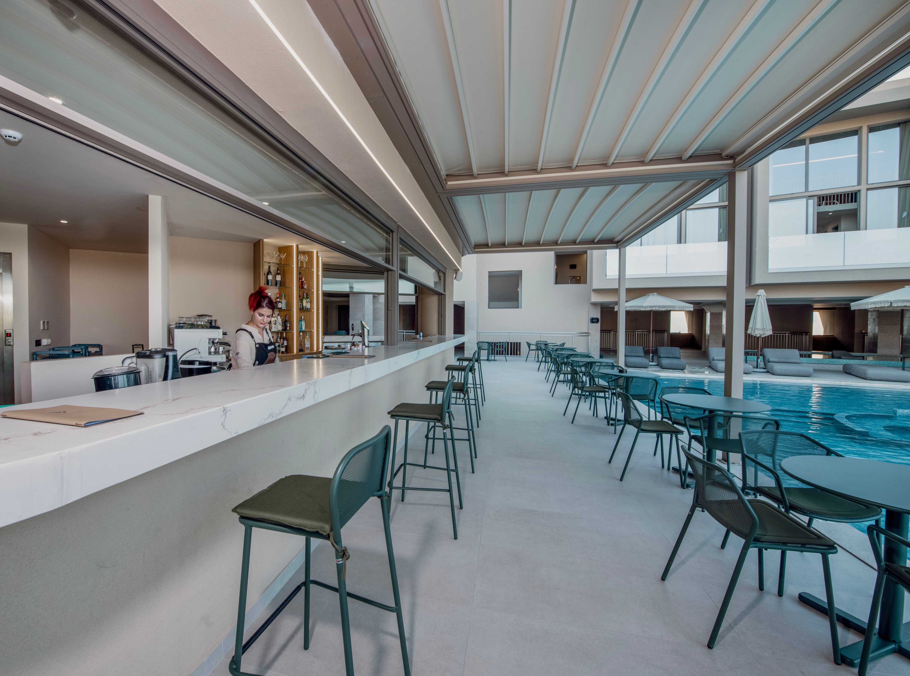 A contemporary open-air bar is situated beside a swimming pool in a resort setting. Green metal barstools and chairs line the long white counter and nearby tables, creating ample seating for guests. A staff member stands behind the bar, with shelves and equipment visible in the background. The space is covered by a sleek pergola-style roof, and the surrounding architecture features clean lines and large windows.