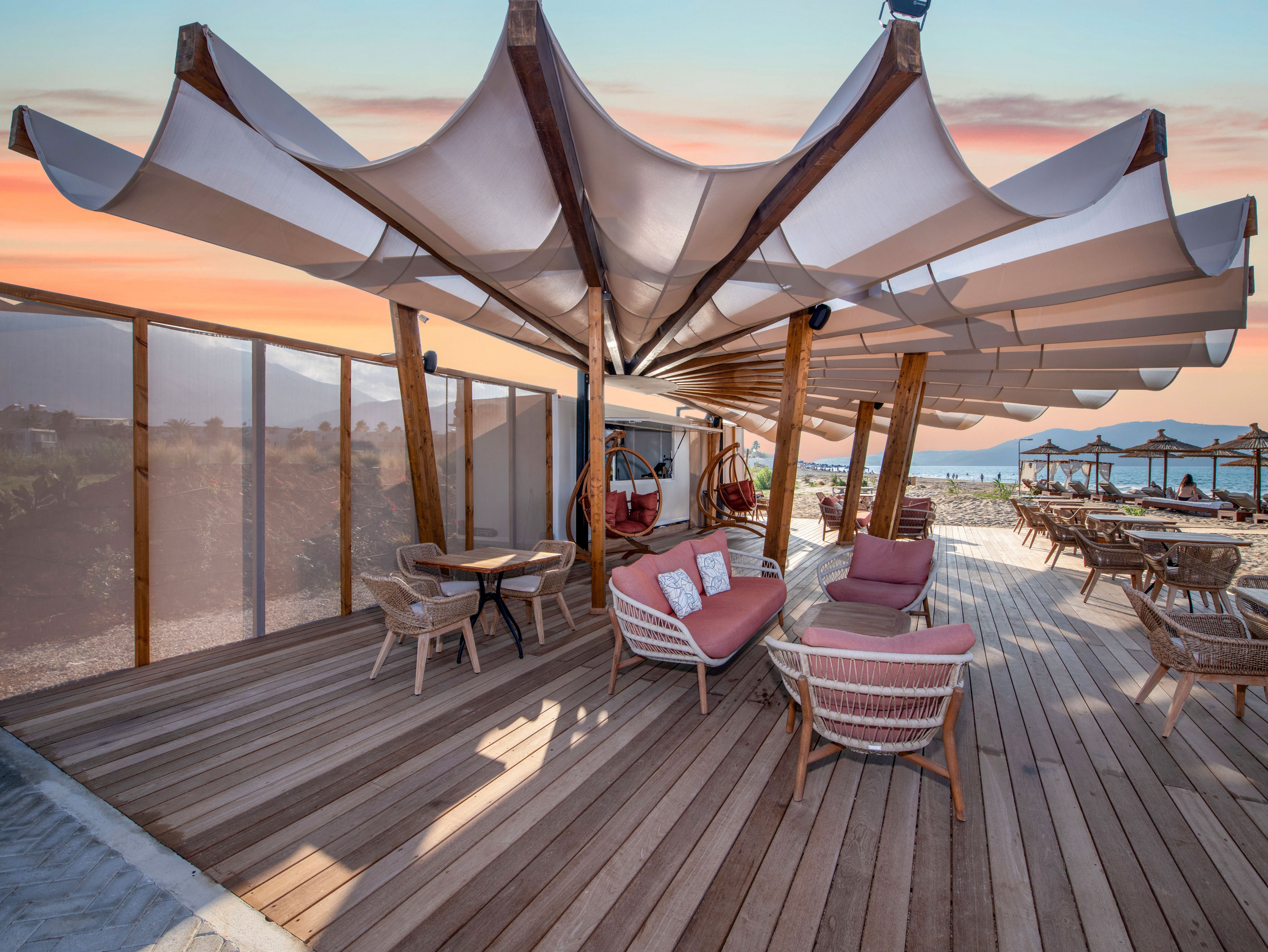 An open-air beachfront lounge features woven chairs and cushioned sofas arranged on a wide wooden deck. Sculptural fabric canopies stretch overhead, creating shade and visual interest against a colorful sunset sky. In the background, additional seating and umbrellas face the sea, suggesting a relaxed coastal hospitality setting. The scene feels inviting and tranquil, ideal for leisure, socializing, or vacation relaxation.