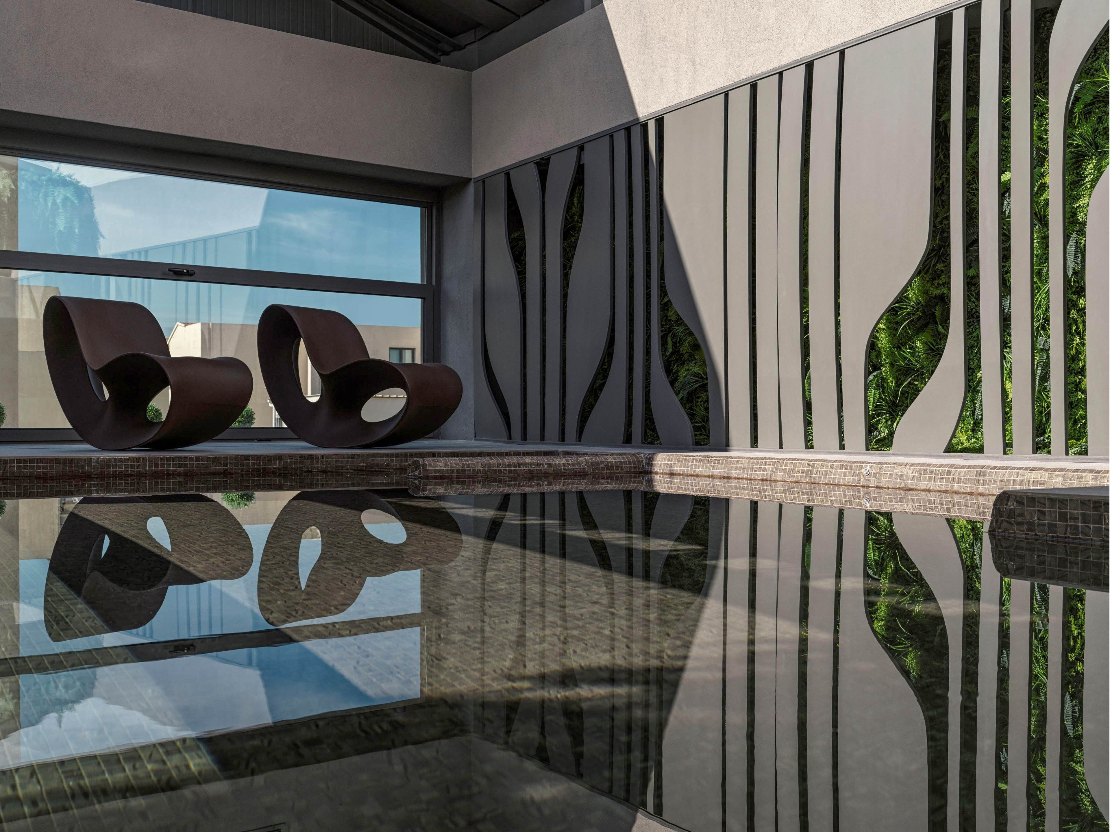 A contemporary indoor pool area features two sculptural brown lounge chairs positioned beside the water. The calm surface of the pool reflects the chairs and surrounding architecture, enhancing the tranquil atmosphere. Vertical decorative panels with organic cutout patterns line one side of the space, while a large horizontal window opens to an exterior view. Soft natural light and muted tones create a minimalist, spa-like setting ideal for relaxation.