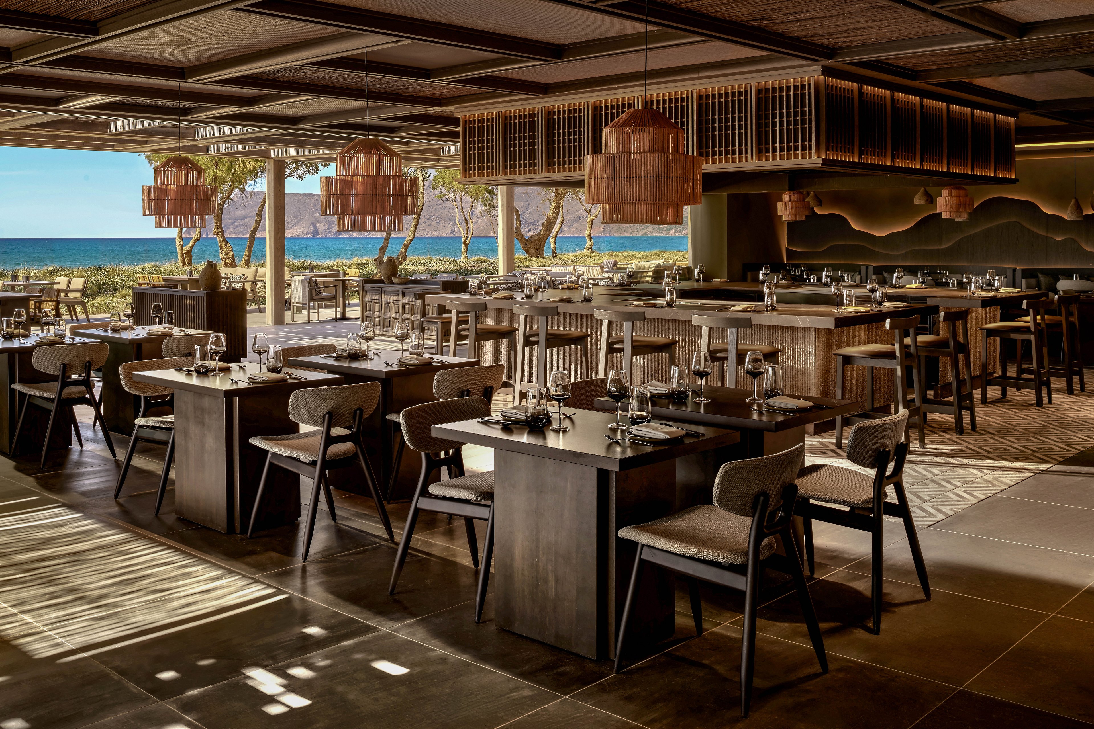 A contemporary restaurant dining room features neatly arranged wooden tables and chairs set with glassware and cutlery. Large windows open to a scenic ocean view with trees and blue water in the background. Warm pendant lights and textured ceiling panels create a cozy yet upscale atmosphere. The space is empty of people, emphasizing the architecture, interior design, and natural light.