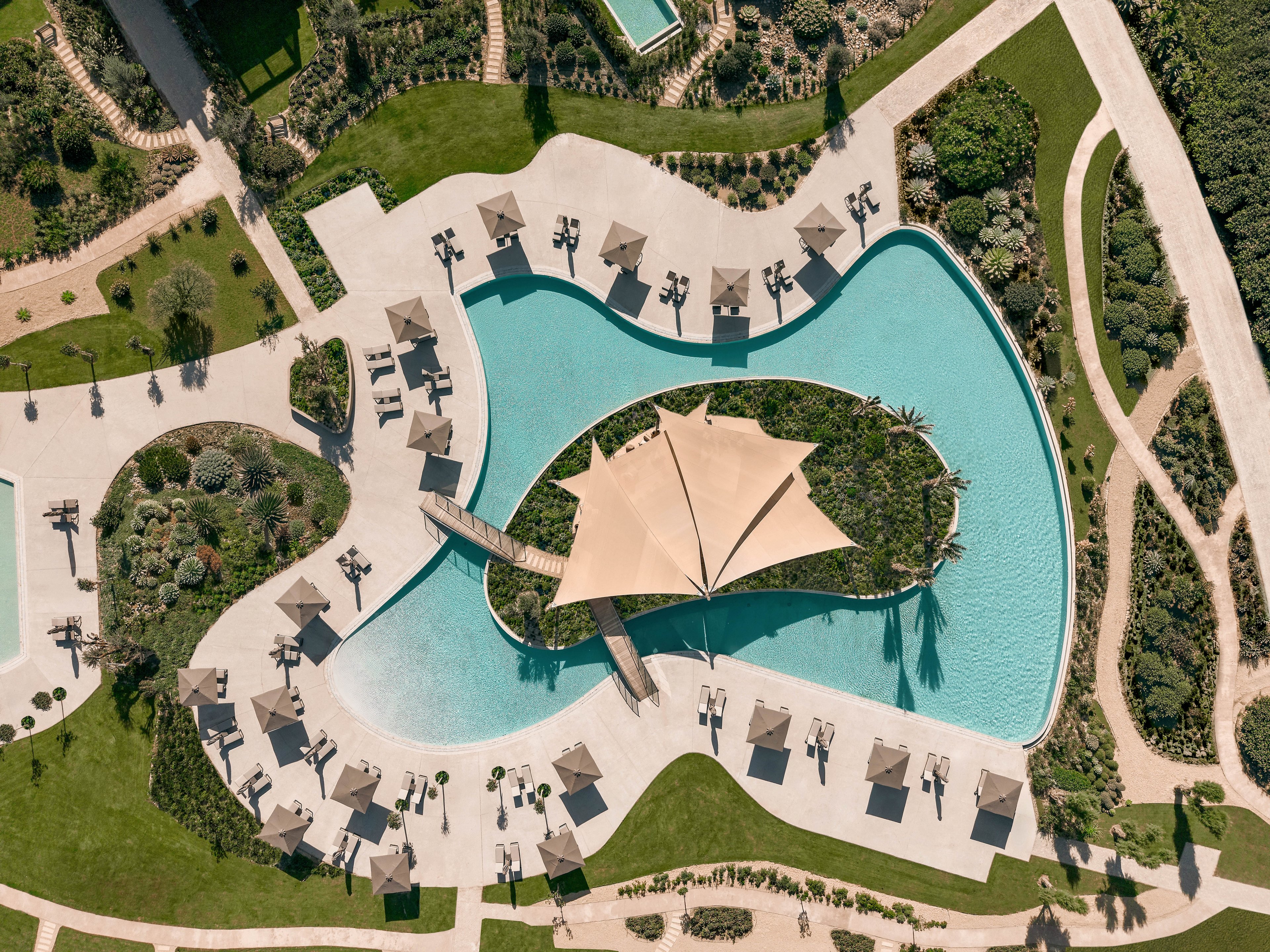 An aerial view showcases a large, organically shaped resort swimming pool surrounded by lush greenery. A central island with plants and a large shade canopy sits in the middle of the pool, connected by a small bridge. Numerous lounge chairs and umbrellas line the curved pool deck, creating a relaxing vacation atmosphere. The surrounding landscaped gardens and pathways emphasize a tropical, luxurious setting.