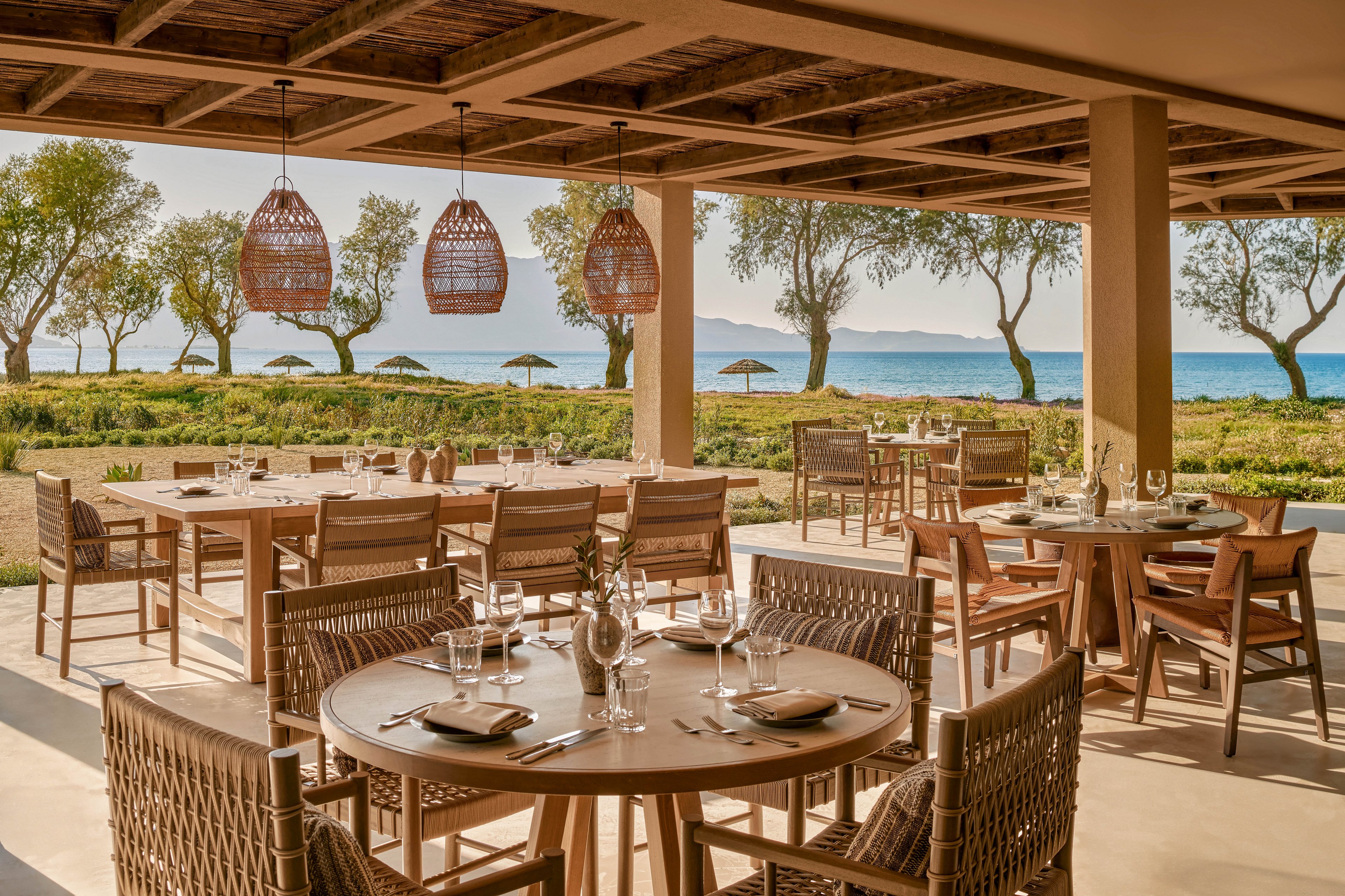 An open-air restaurant terrace features neatly set wooden tables and woven chairs overlooking a scenic beachfront. Glassware, plates, and cutlery are arranged on the tables, suggesting a relaxed dining experience. The covered patio ceiling with hanging wicker lights frames a view of trees, umbrellas, and calm blue water in the distance. Warm natural tones and soft sunlight create an inviting, tranquil atmosphere ideal for seaside meals.
