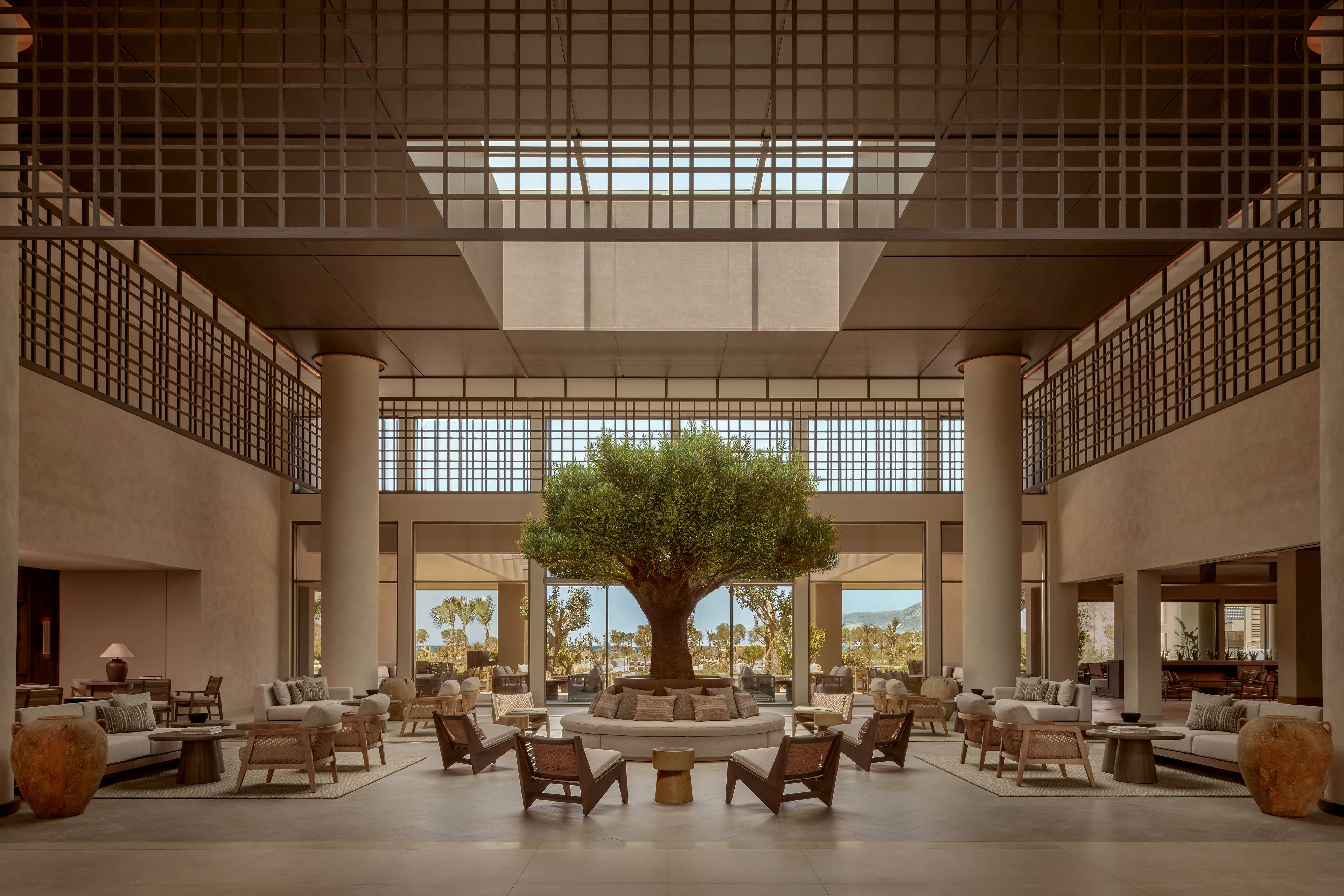 A spacious, contemporary hotel lobby features a large tree planted in a circular seating area at its center. Neutral-toned sofas and armchairs are arranged symmetrically around the tree, creating an inviting lounge space. Tall columns, grid-like ceiling structures, and expansive windows frame views of palm trees and distant mountains. Warm lighting and natural materials contribute to a calm, resort-like atmosphere.