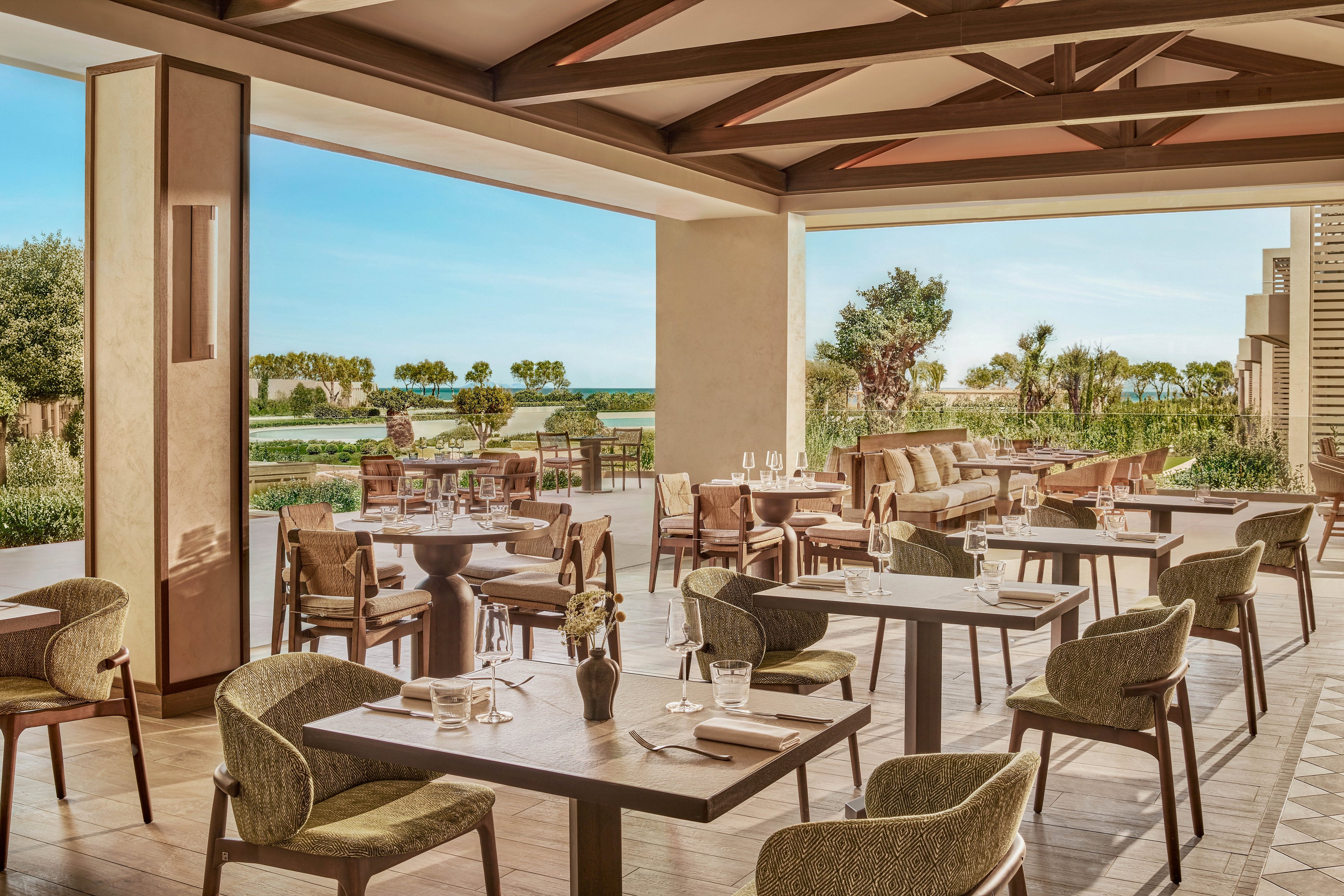 An elegant open-air restaurant features neatly arranged wooden tables and cushioned chairs under a covered terrace. Place settings with glassware and cutlery are prepared, suggesting a refined dining experience. Beyond the seating area, manicured lawns, trees, and a distant view of the sea create a serene coastal backdrop. Warm neutral tones and natural materials give the space a relaxed, upscale resort ambiance.