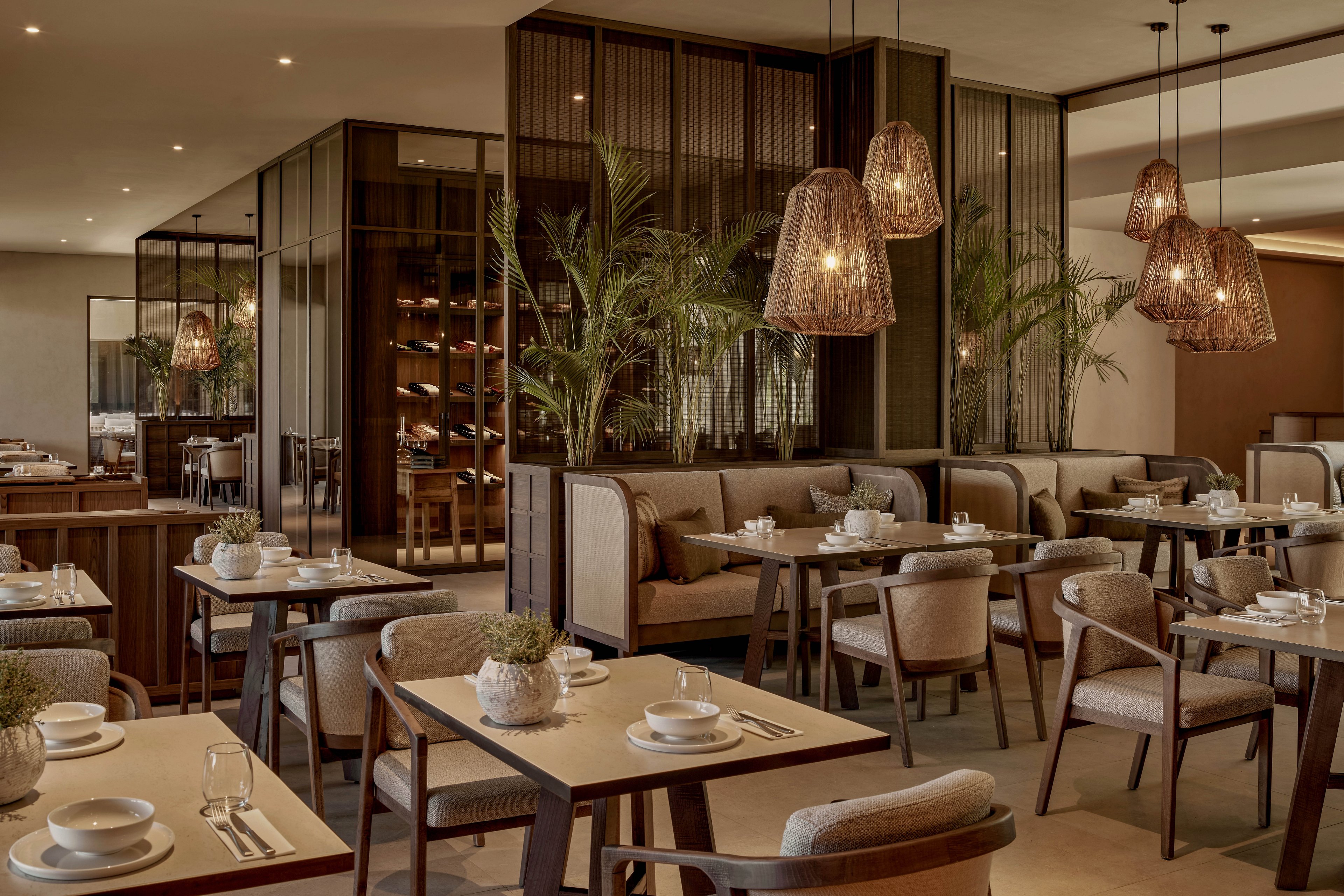 A contemporary restaurant dining area features neatly arranged tables set with white dishes and cups. Soft upholstered chairs and high-backed booths create an inviting, comfortable seating layout. Warm pendant lights with woven shades hang from the ceiling, casting a cozy glow over the space. Tall plants and wooden partitions add natural accents and structure to the sophisticated, neutral-toned interior.