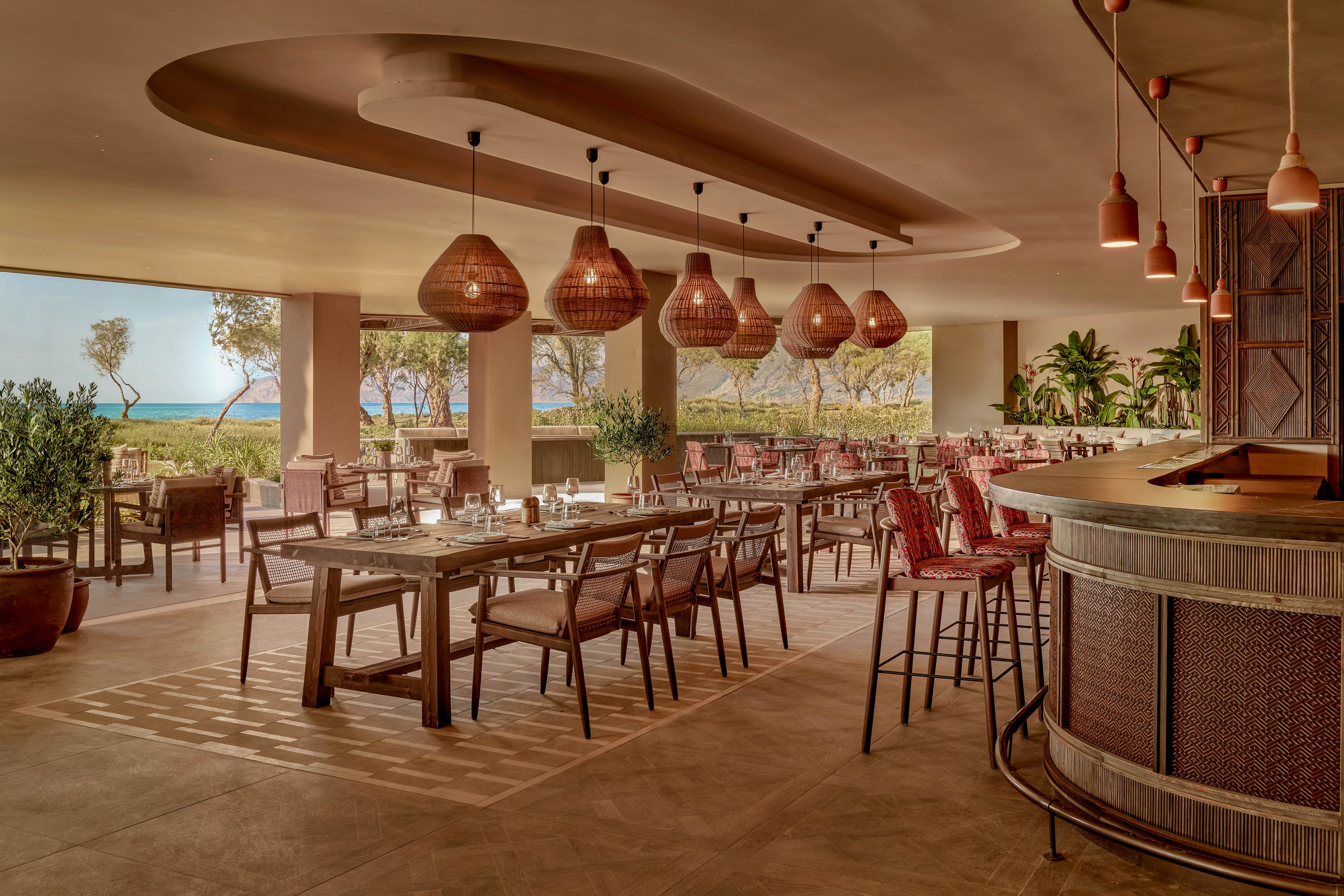 Spacious restaurant interior features wooden tables and chairs arranged neatly under sculptural pendant lights. Large open windows reveal a coastal landscape with trees and a view of the sea in the background. The bar area curves along one side, complemented by earthy tones, woven textures, and potted plants. Soft, warm lighting and natural materials create a relaxed, inviting dining atmosphere.