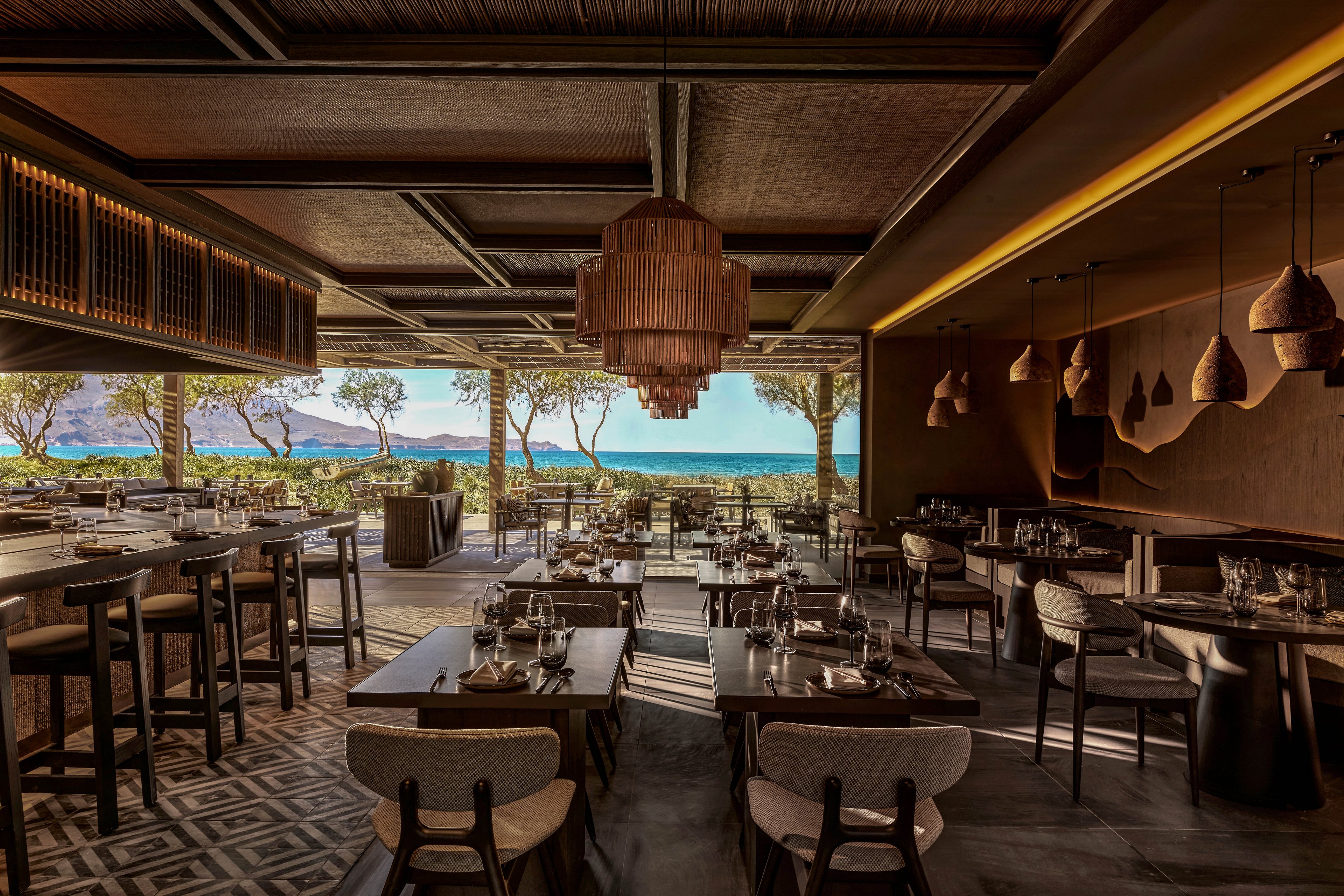 An elegant, contemporary restaurant interior opens onto a terrace with a scenic ocean and mountain backdrop. Dark wooden tables and high-top seating are neatly arranged, each set with glassware and table settings. Warm pendant lighting and textured ceiling details create a cozy, sophisticated atmosphere. Large open windows frame trees and the glowing horizon, blending indoor comfort with an outdoor coastal landscape.