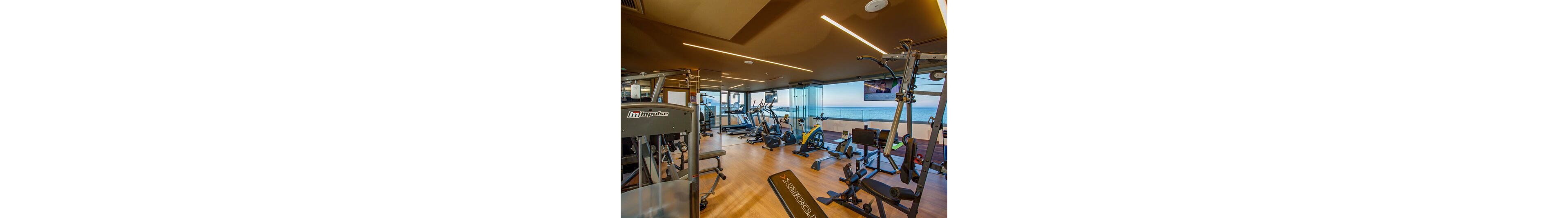 A contemporary fitness room is equipped with various exercise machines, including treadmills, stationary bikes, weight benches, and strength-training equipment. Large floor-to-ceiling windows reveal a scenic ocean view and an outdoor terrace, creating an open and airy atmosphere. Warm ceiling lighting and wood-style flooring contribute to a clean, modern design. The space appears organized and inviting for guests seeking a workout with a coastal backdrop.