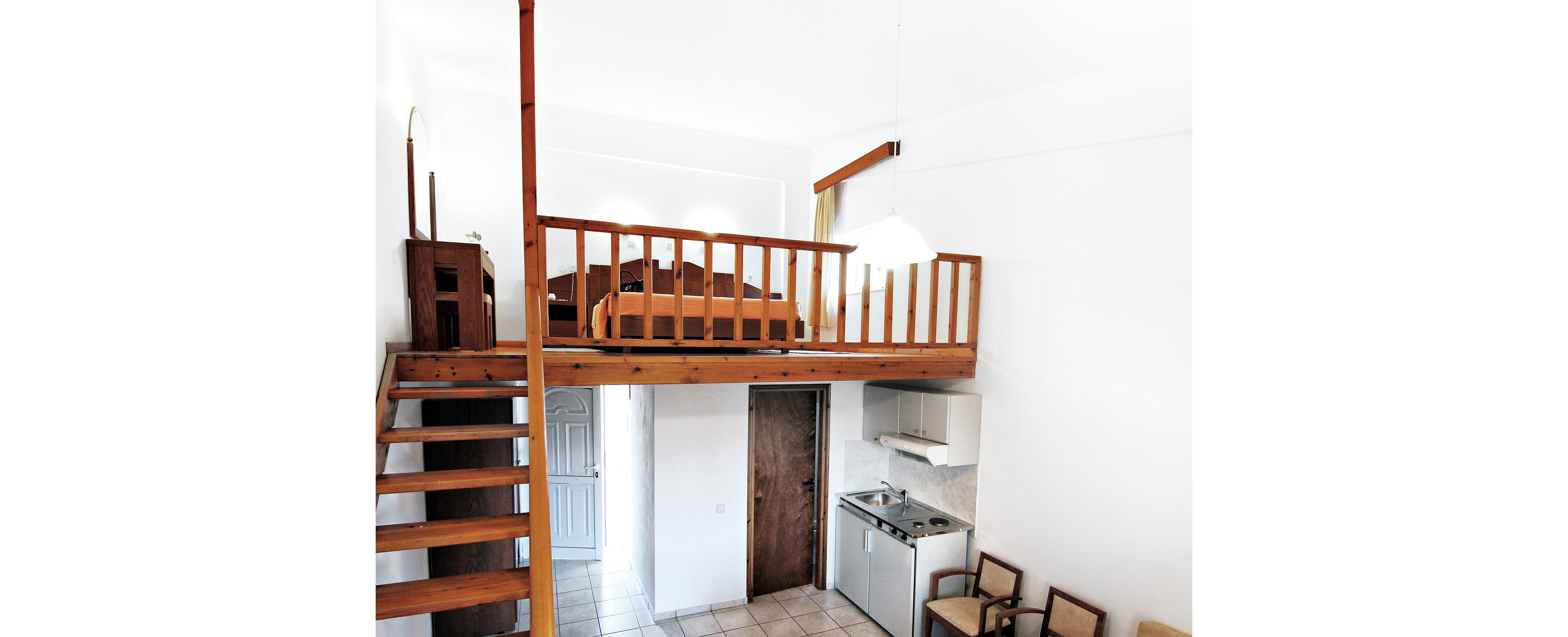 A compact loft apartment features a wooden mezzanine level with a bed and railing above a small kitchenette. Wooden stairs lead up to the sleeping area, while a tiled floor and white walls enhance the bright, airy feel. Below the loft, a simple kitchen with sink, cabinets, and mini stove sits beside a wooden door and a row of chairs. A hanging light fixture and natural light from an unseen window illuminate the minimalist interior.